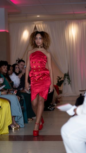 Model struts down catwalk in red satin mini dress with gathered details at fashion show.
