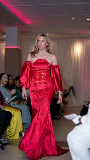 Model presents bright red satin dress with puff sleeves and gold button details on fashion runway.