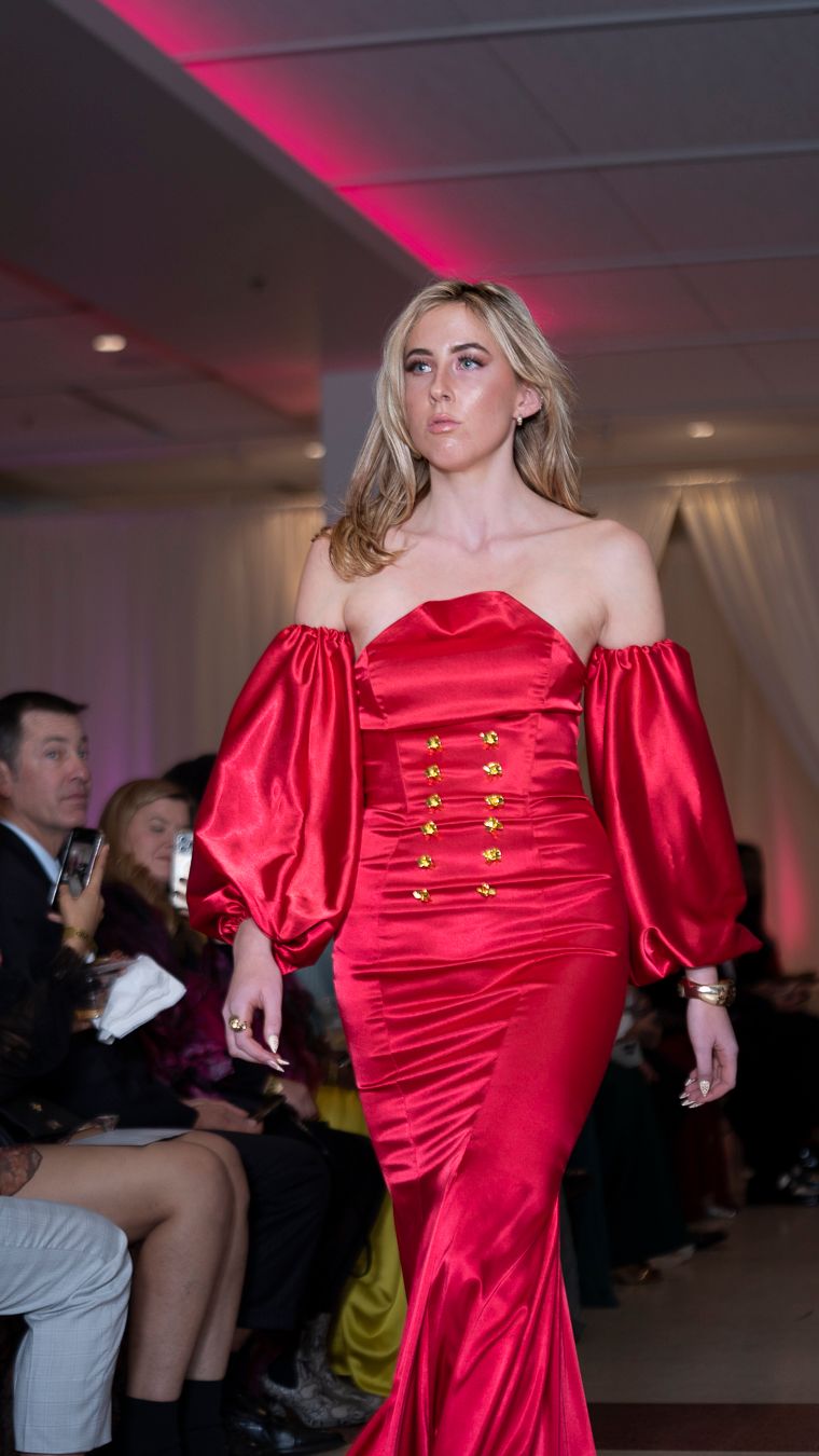 Red satin off-shoulder gown with puff sleeves and gold buttons on fashion catwalk.