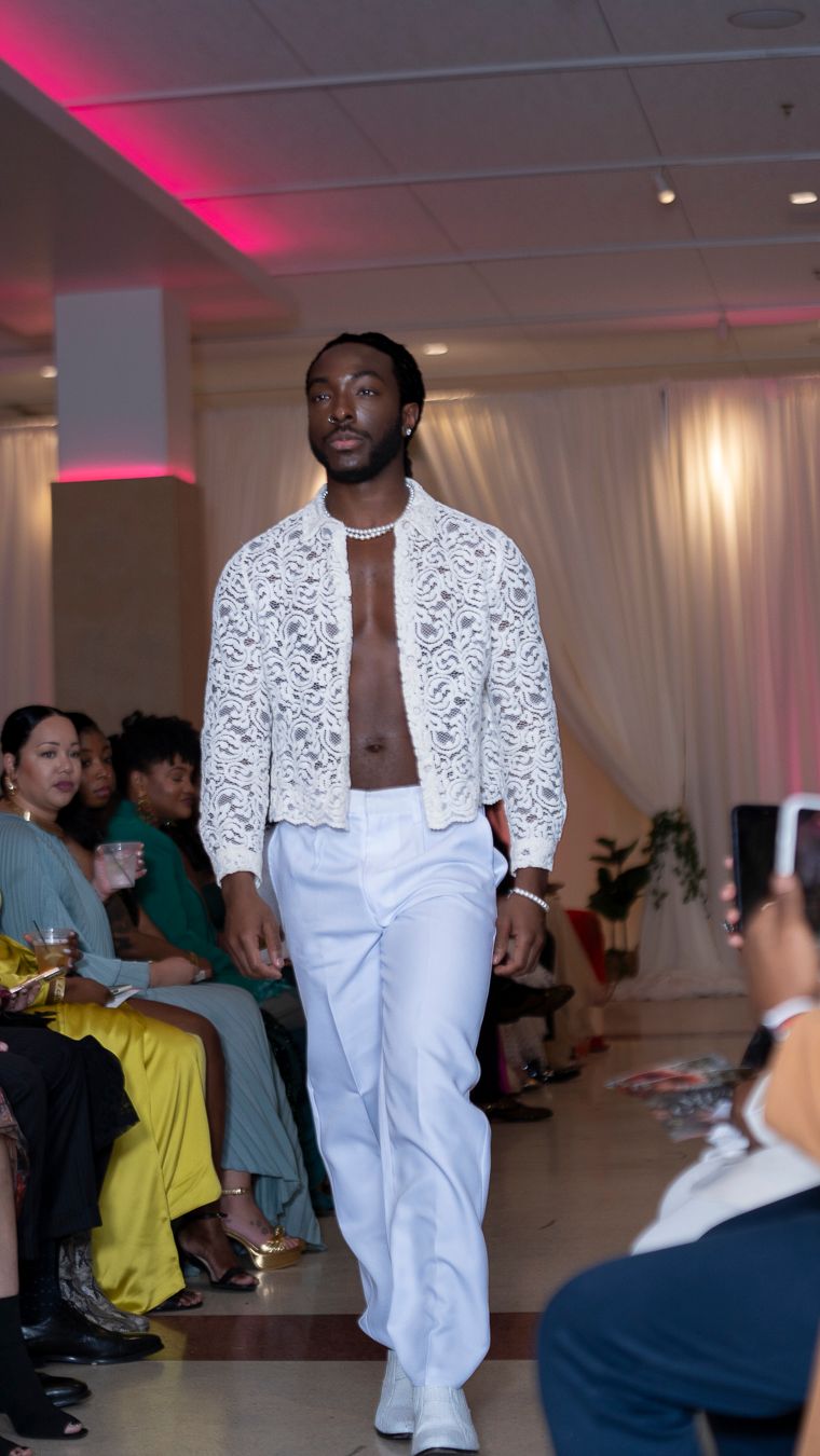 Fashion model walks runway in white embellished jacket and pants ensemble at indoor fashion show with pink lighting.