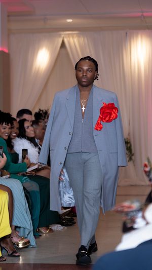 Model walks runway in light gray suit with red flower accent during fashion show with audience seated along sides.
