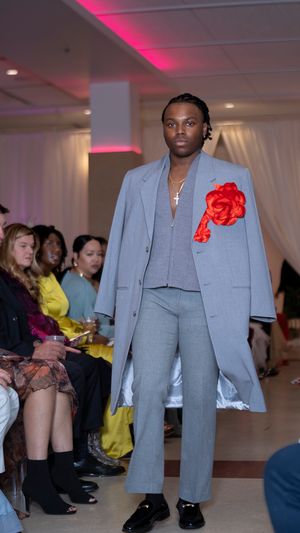 Model walks runway in gray suit with red flower accent during fashion show with pink lighting.