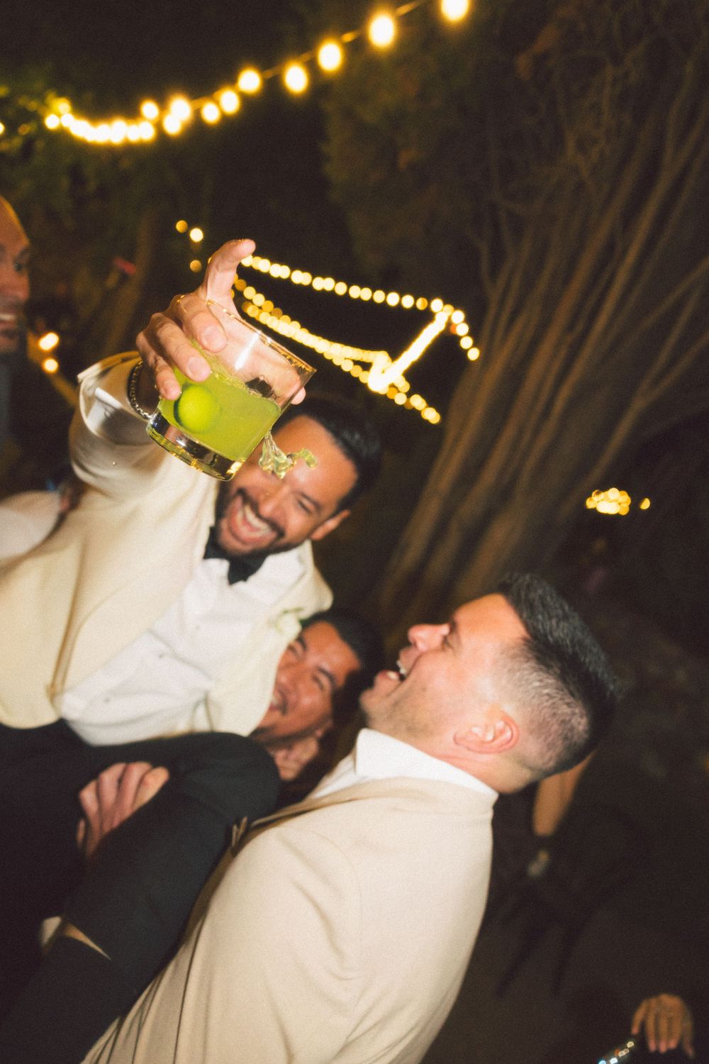 A person in white clothing drinks from a bottle under string lights at an outdoor nighttime event.