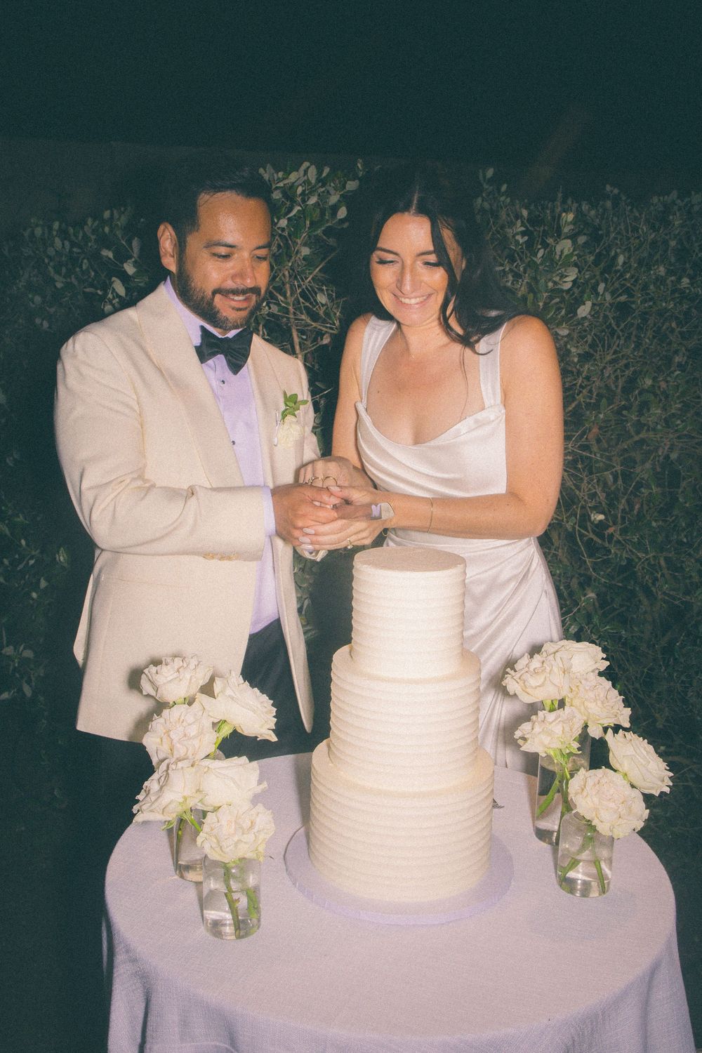 A couple cuts their elegant white wedding cake together while surrounded by floral arrangements in glass vases.