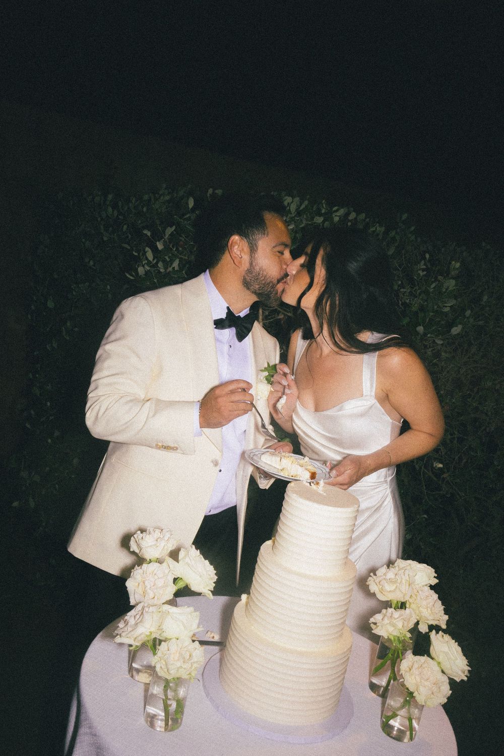 A couple shares a romantic moment while cutting their elegant white wedding cake surrounded by floral arrangements.
