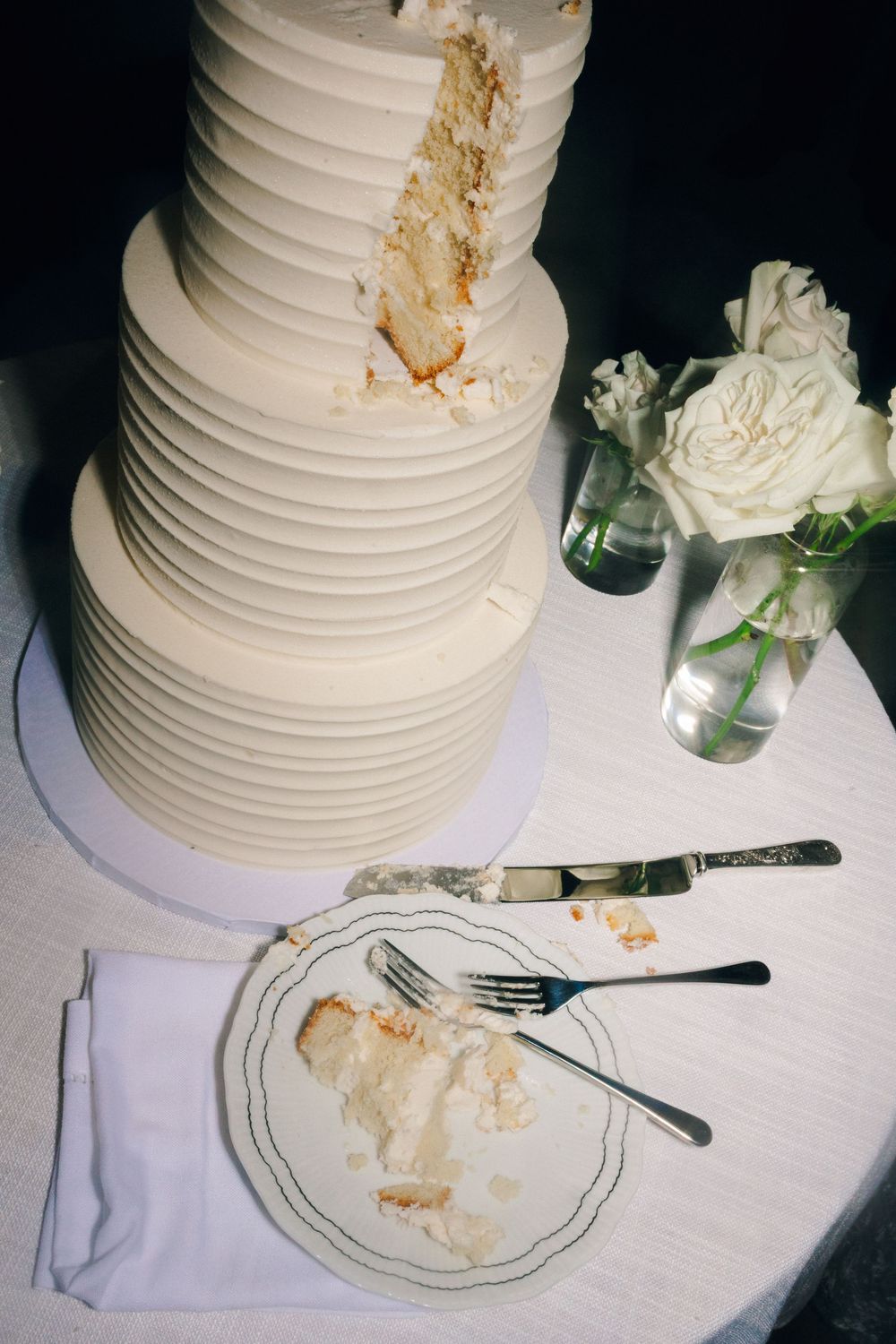 A white tiered wedding cake with horizontal ribbing sits next to a plate with crumbs and a vase of white roses.