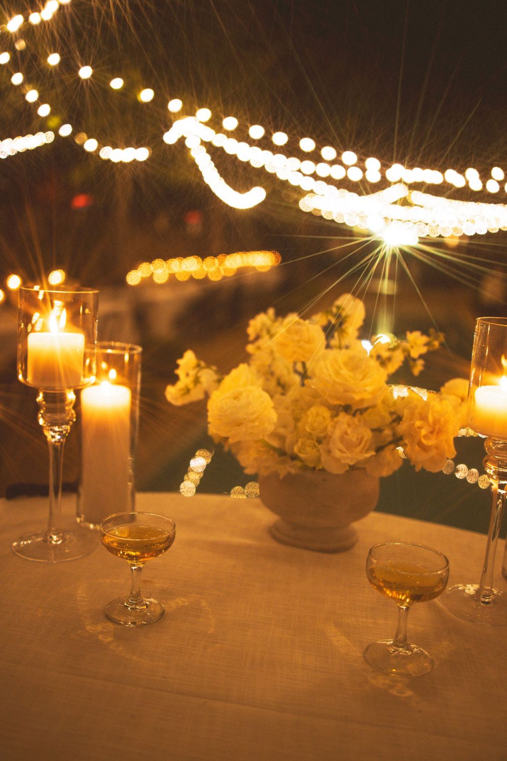 String lights illuminate an elegant wedding reception table with glowing candles and yellow floral centerpieces.