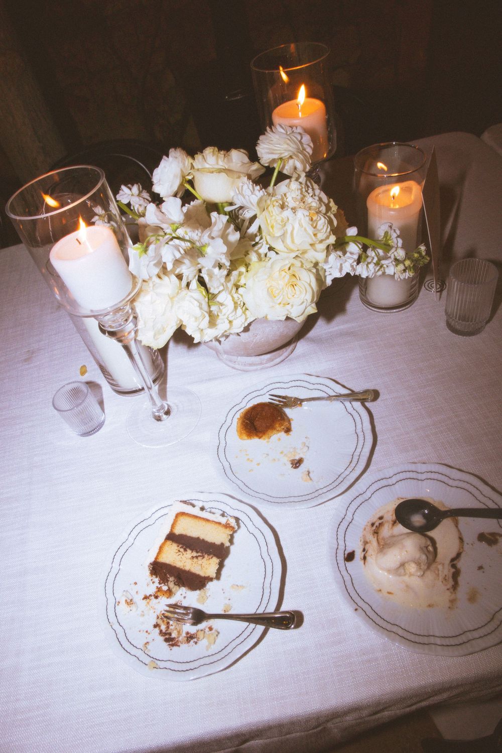 Romantic table setting with white roses, lit candles, and partially eaten desserts on elegant plates.