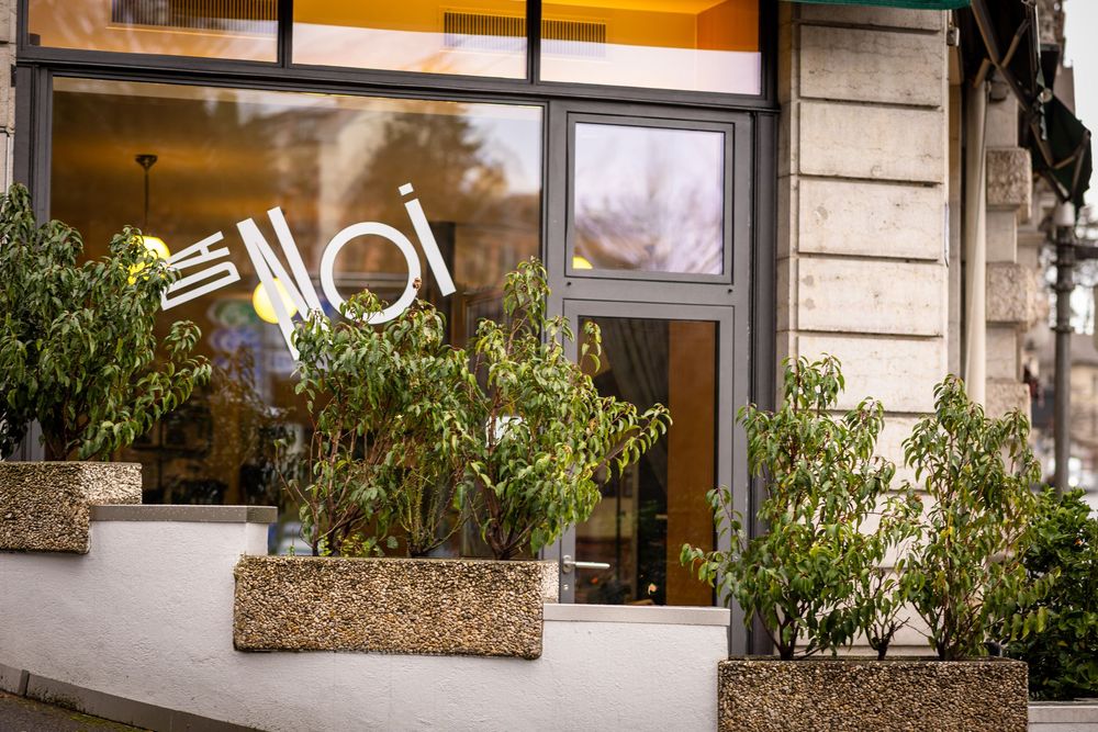 Modern restaurant exterior with glass windows, potted plants, and stone facade illuminated at dusk.