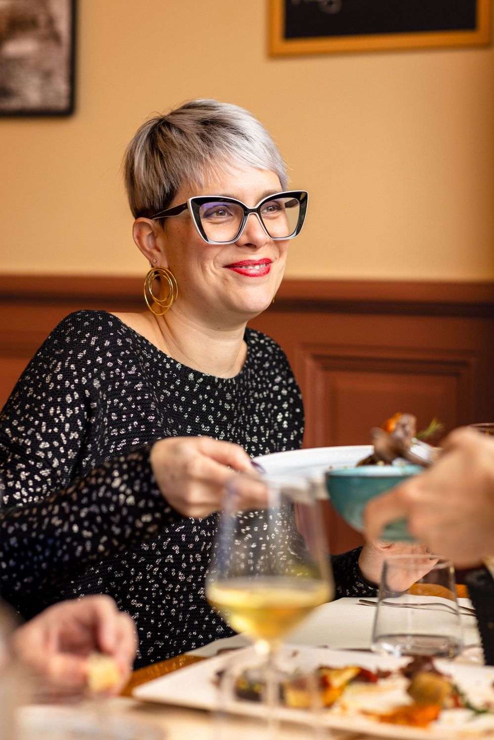 A person with glasses and blonde hair wearing a polka dot dress enjoys a meal and drinks at a restaurant table.