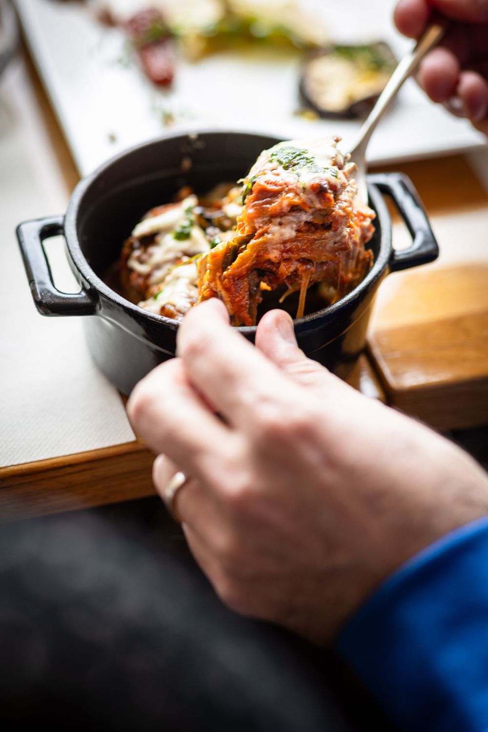 Hands using utensils to serve a steaming portion of pasta with sauce from a black cast iron pot.