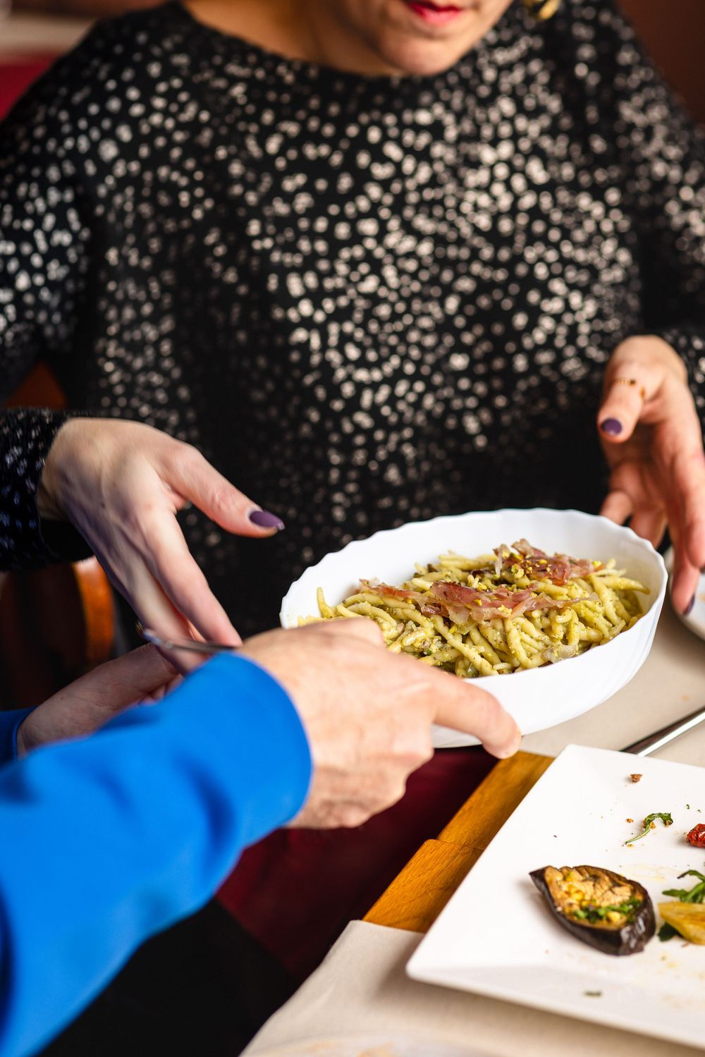 Bowl of pasta being served at restaurant table with grilled vegetables on side plate.