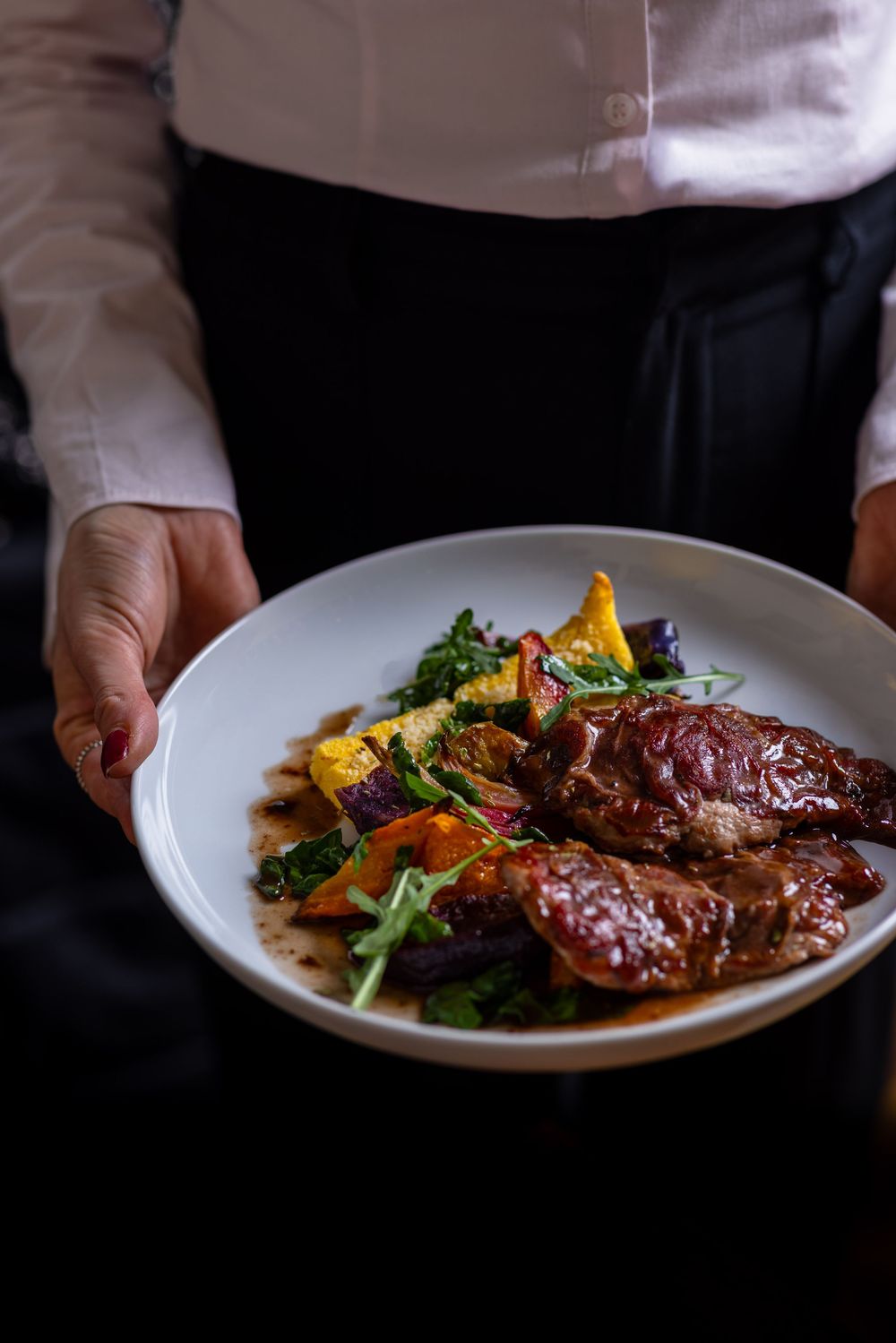 Hands holding white plate with glazed meat dish garnished with colorful vegetables and microgreens in dark setting.