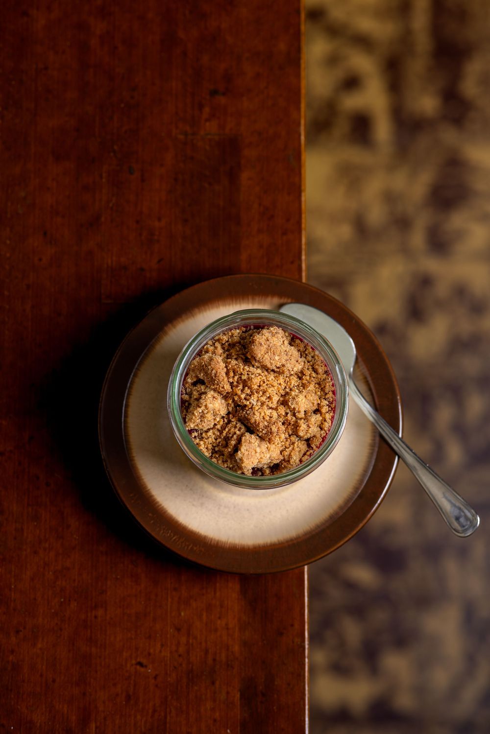 A bowl of brown sugar or crumble topping sits on a dark wooden surface, photographed from above with dramatic lighting.