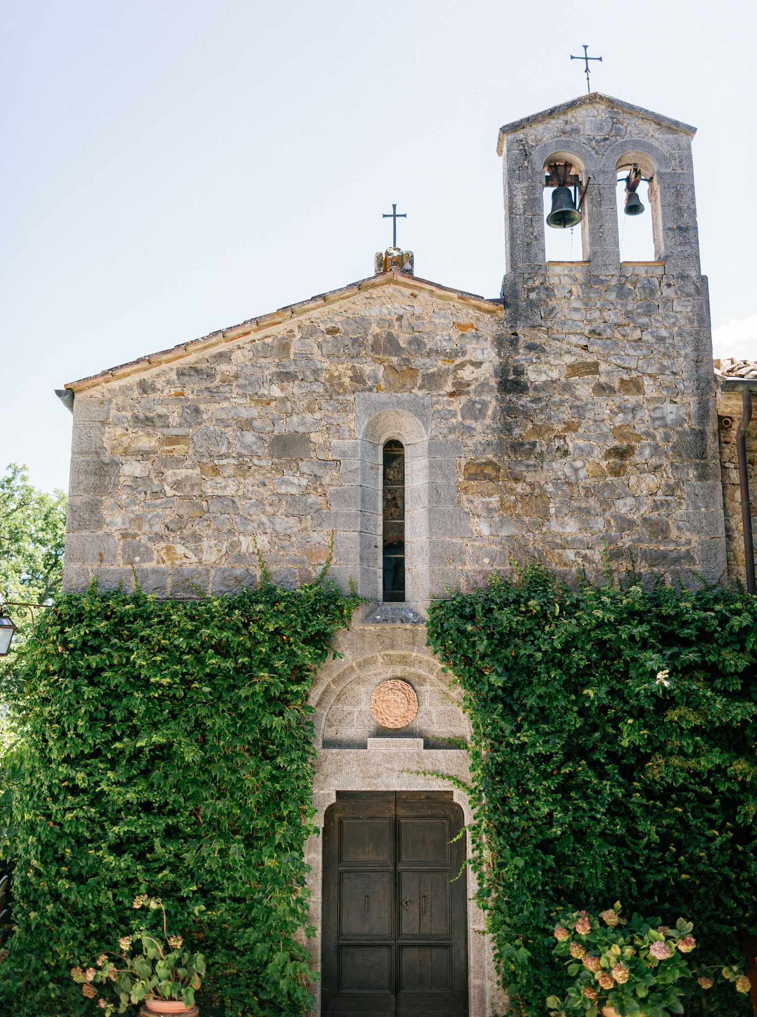 A rustic stone church facade with bell tower and dark wooden doors surrounded by lush green climbing vines.