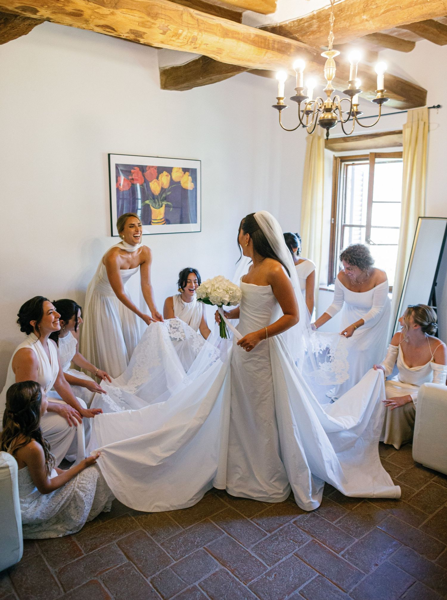A group helps fluff and arrange a white wedding dress in a rustic room with exposed wooden beams and chandeliers.