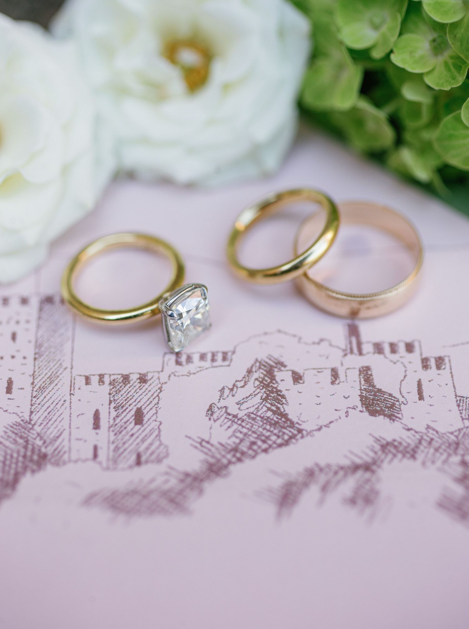 Two gold wedding rings rest on a delicate pink surface with sketched architectural drawings in the background.