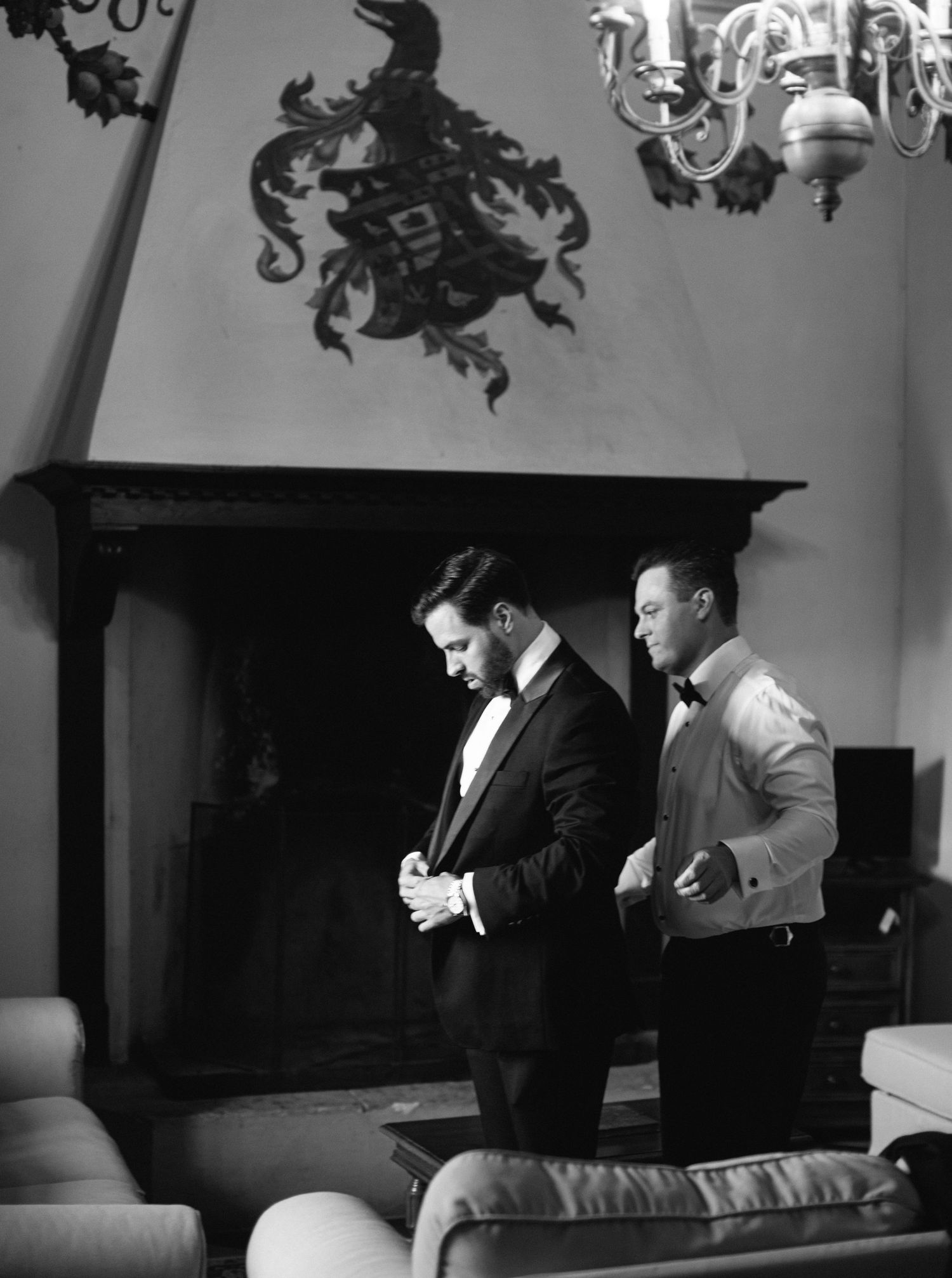Two men in formal attire adjusting their suits while standing in an elegant room with a chandelier and decorative wall crest.