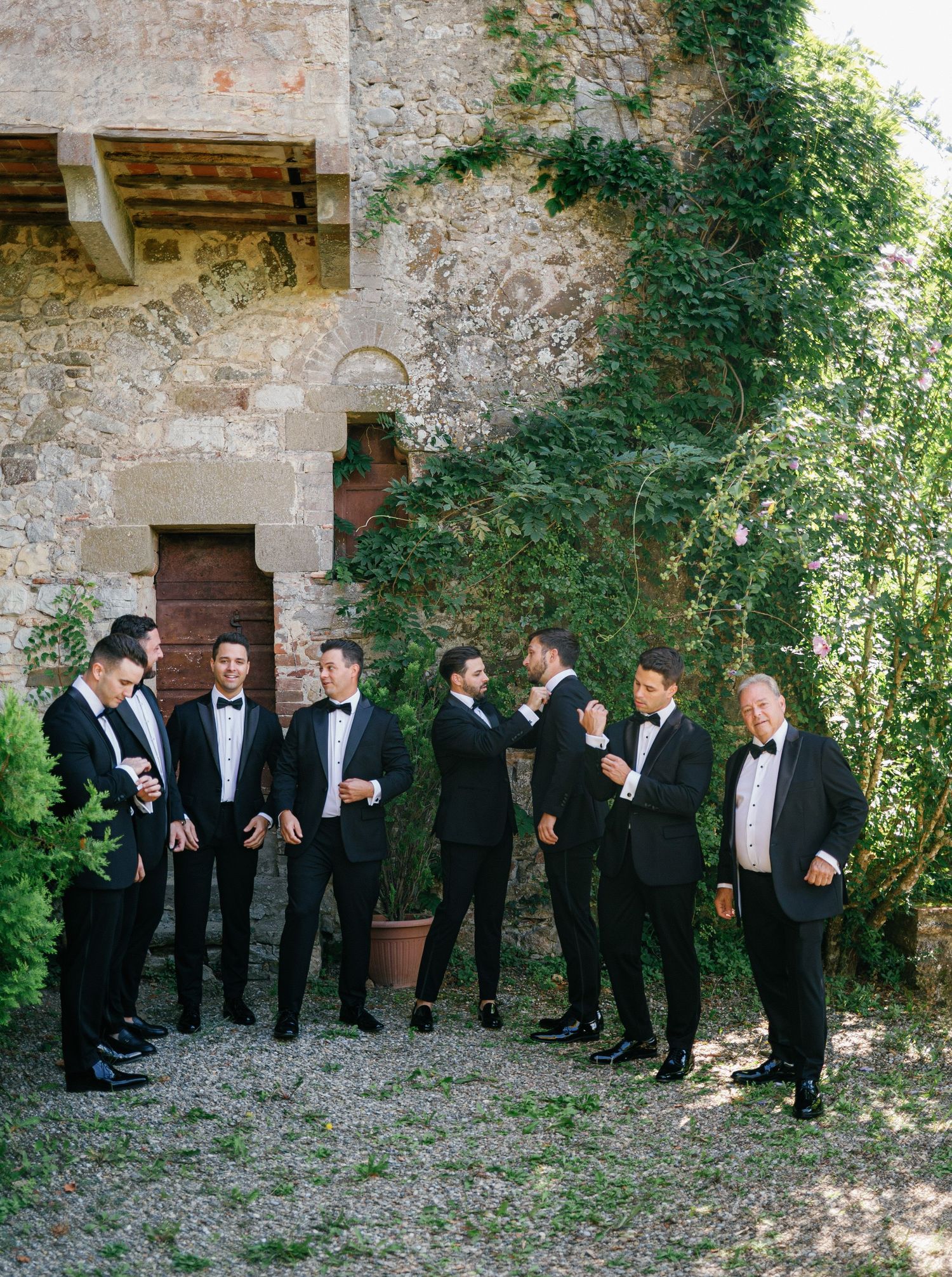 A group in formal black tuxedos gathers on gravel ground against an old stone wall with climbing vines.