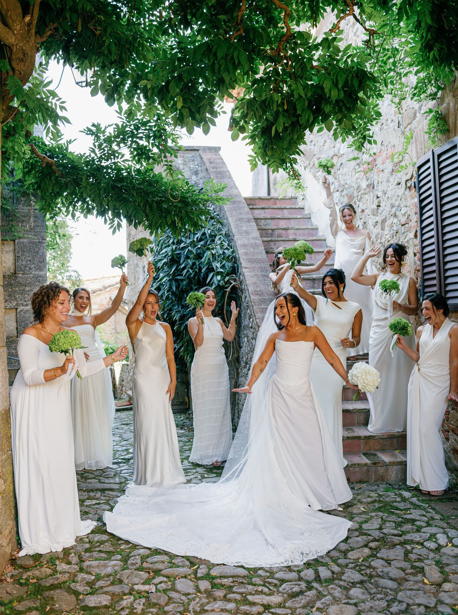 A group in white dresses poses joyfully together on a stone patio surrounded by lush greenery and trees.