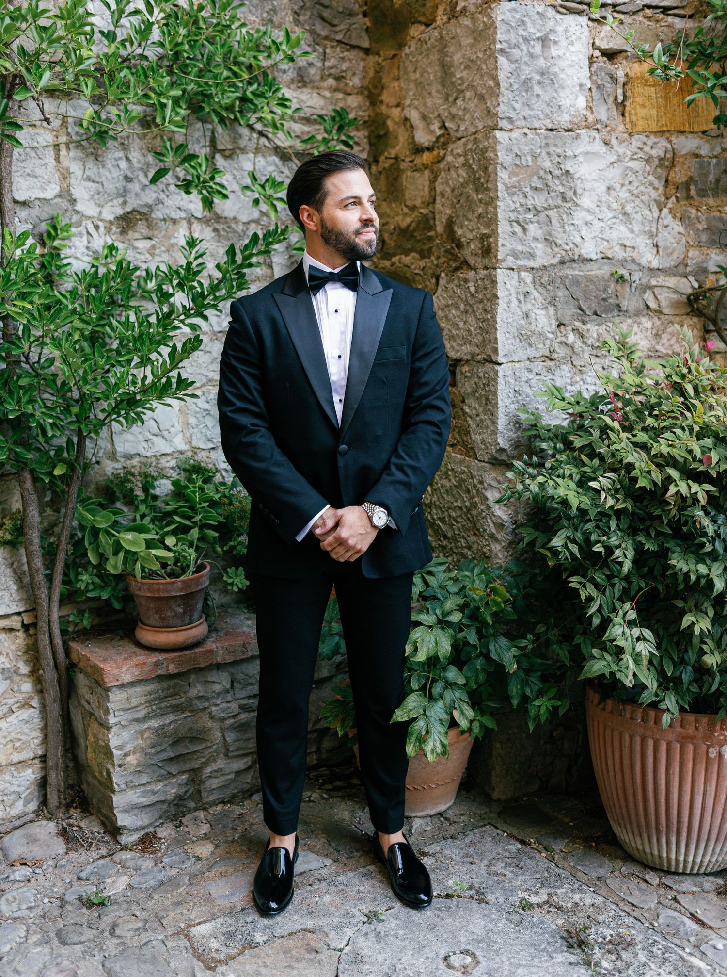 A groom in a classic black tuxedo poses against a rustic stone wall with potted plants in an outdoor setting.