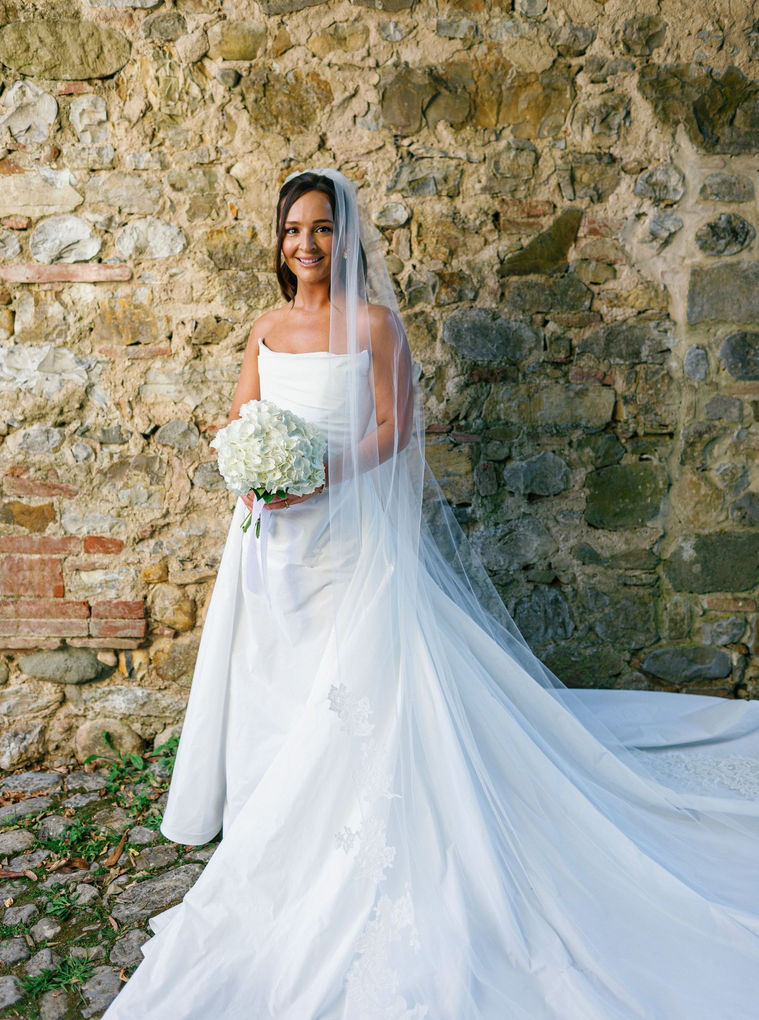 A bride in a flowing white gown with long veil holds a white bouquet against a rustic stone wall backdrop.