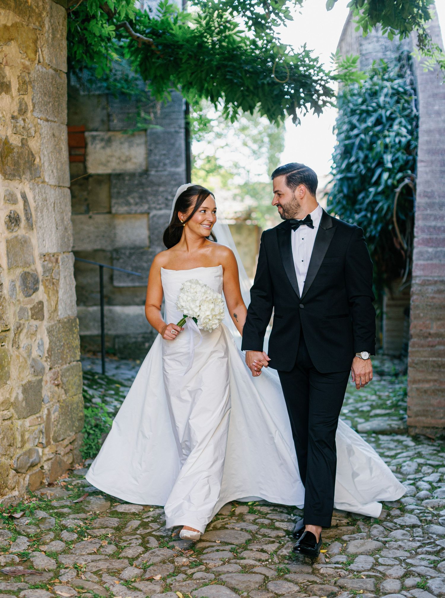 A couple in wedding attire walks along a cobblestone path framed by rustic stone walls and lush greenery.