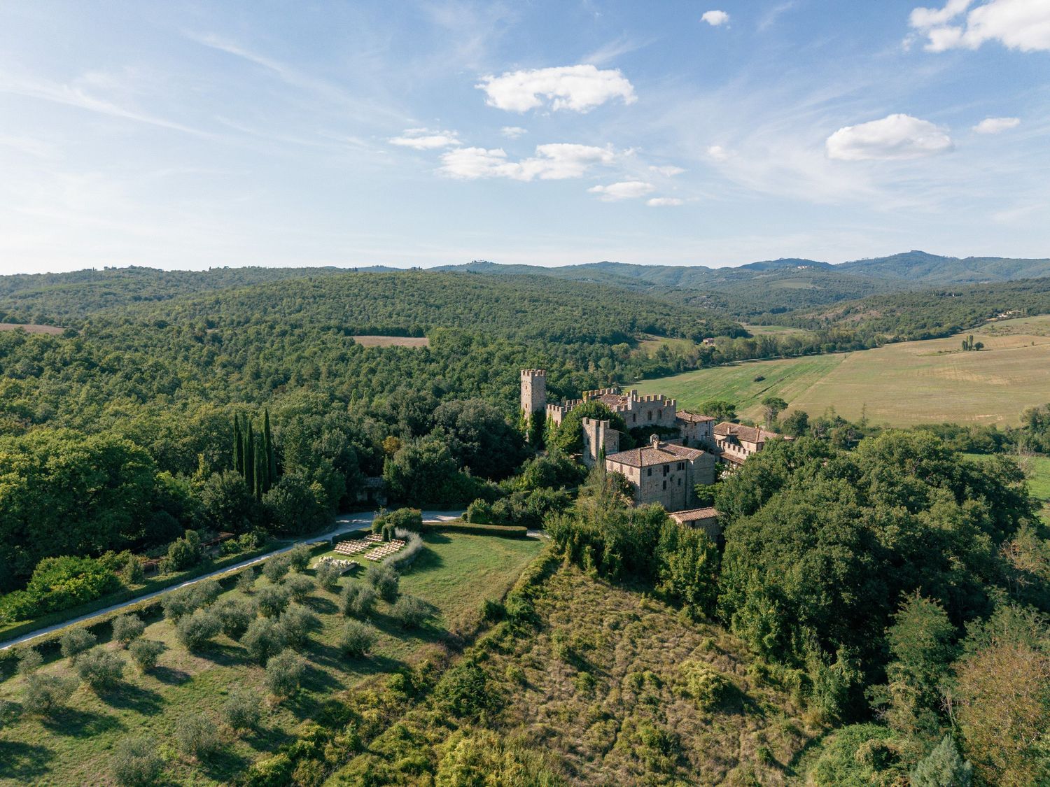 Panoramic aerial view of rolling Tuscan hills with scattered villas and cypress trees under a bright blue summer sky.