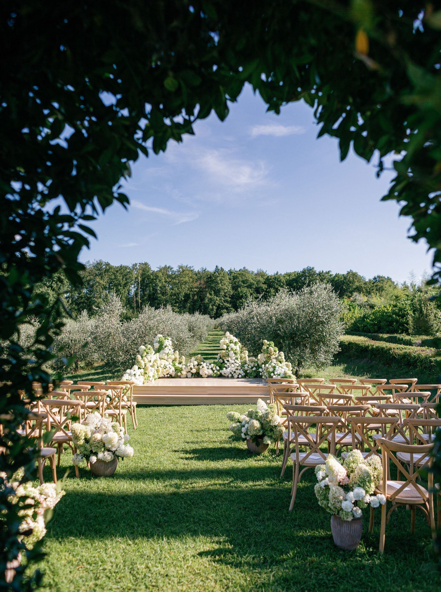 Rustic wooden cross-back chairs arranged in rows on a grassy lawn for an outdoor wedding ceremony with white floral decorations.