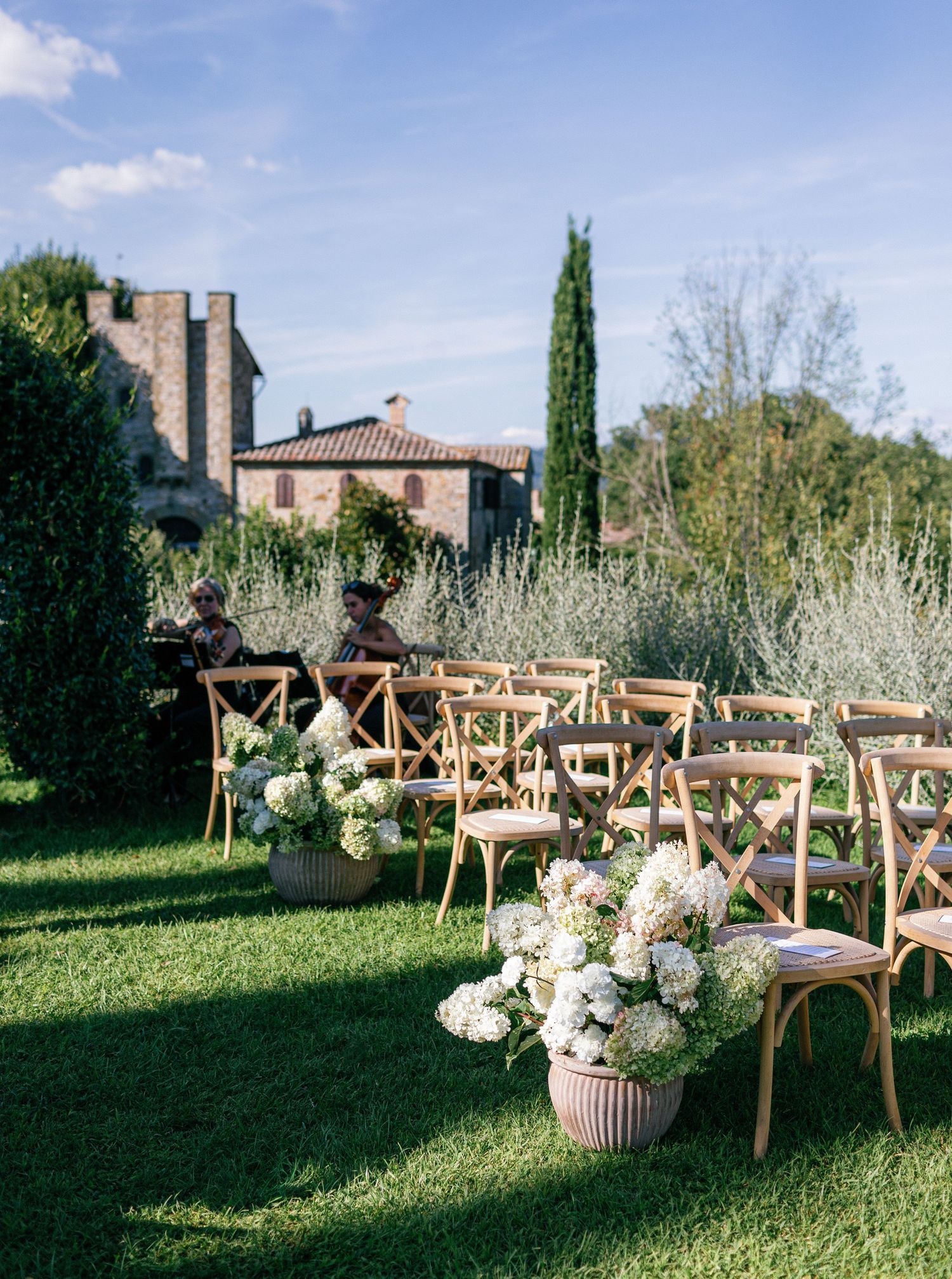 Rustic wooden cross-back chairs arranged in rows on a lush green lawn for an outdoor garden wedding ceremony.