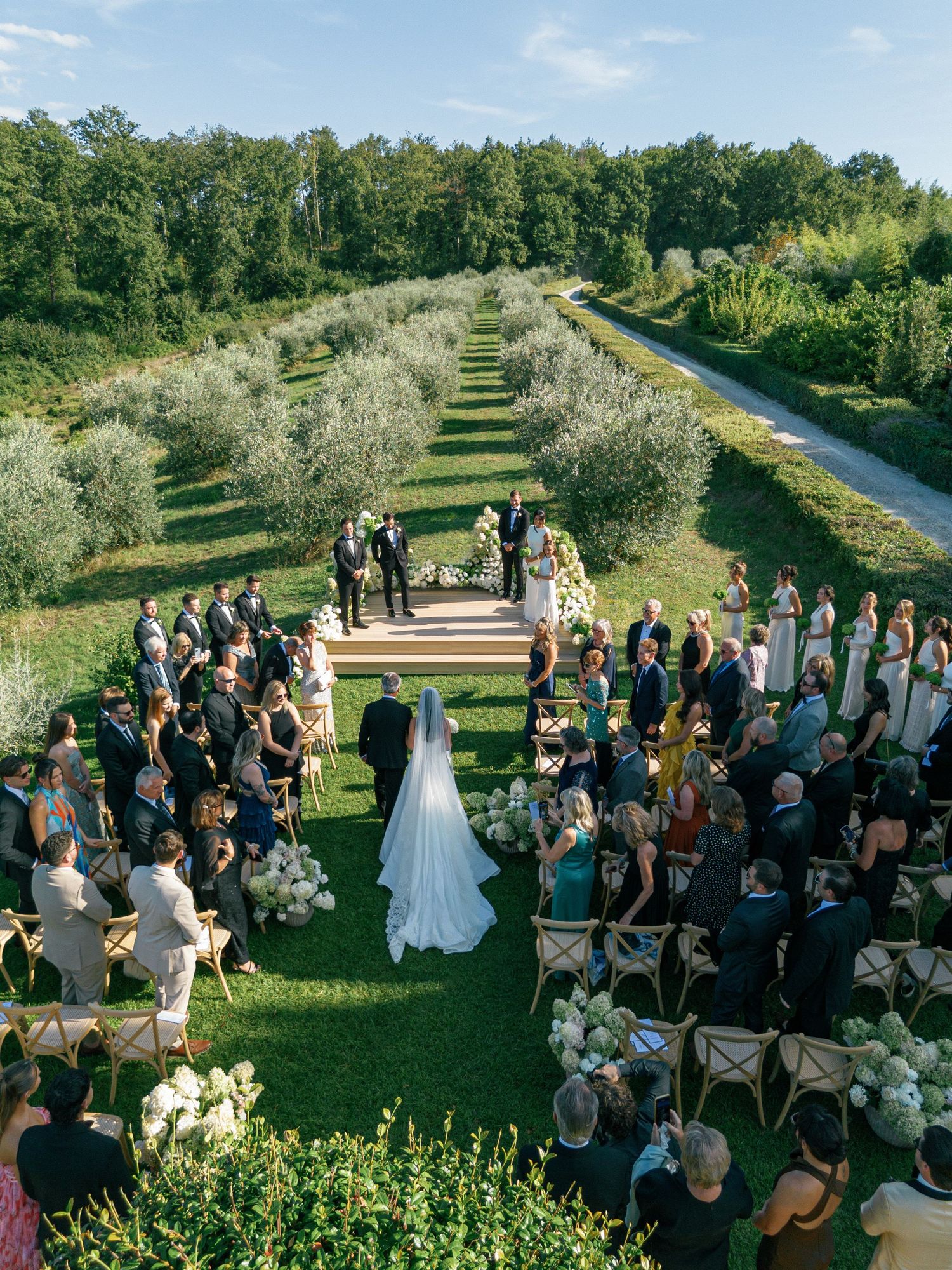 Outdoor wedding ceremony on a grassy hillside with guests seated in rows as the sun casts golden light through trees.