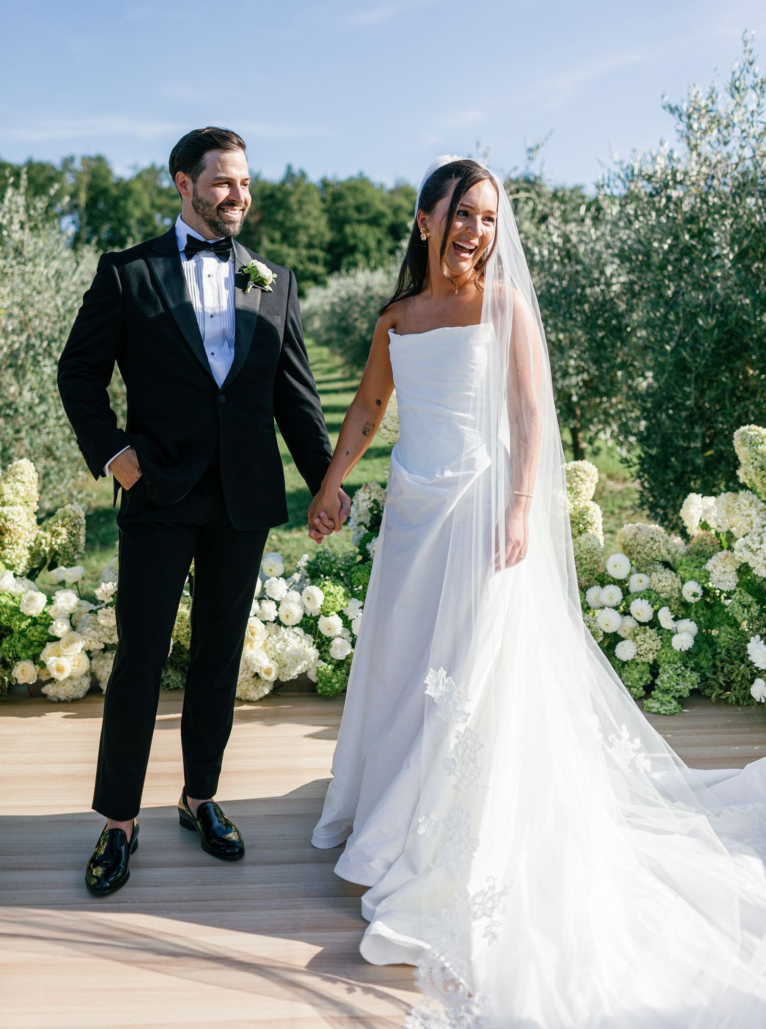 A couple in wedding attire walking together on a wooden deck surrounded by white floral arrangements.