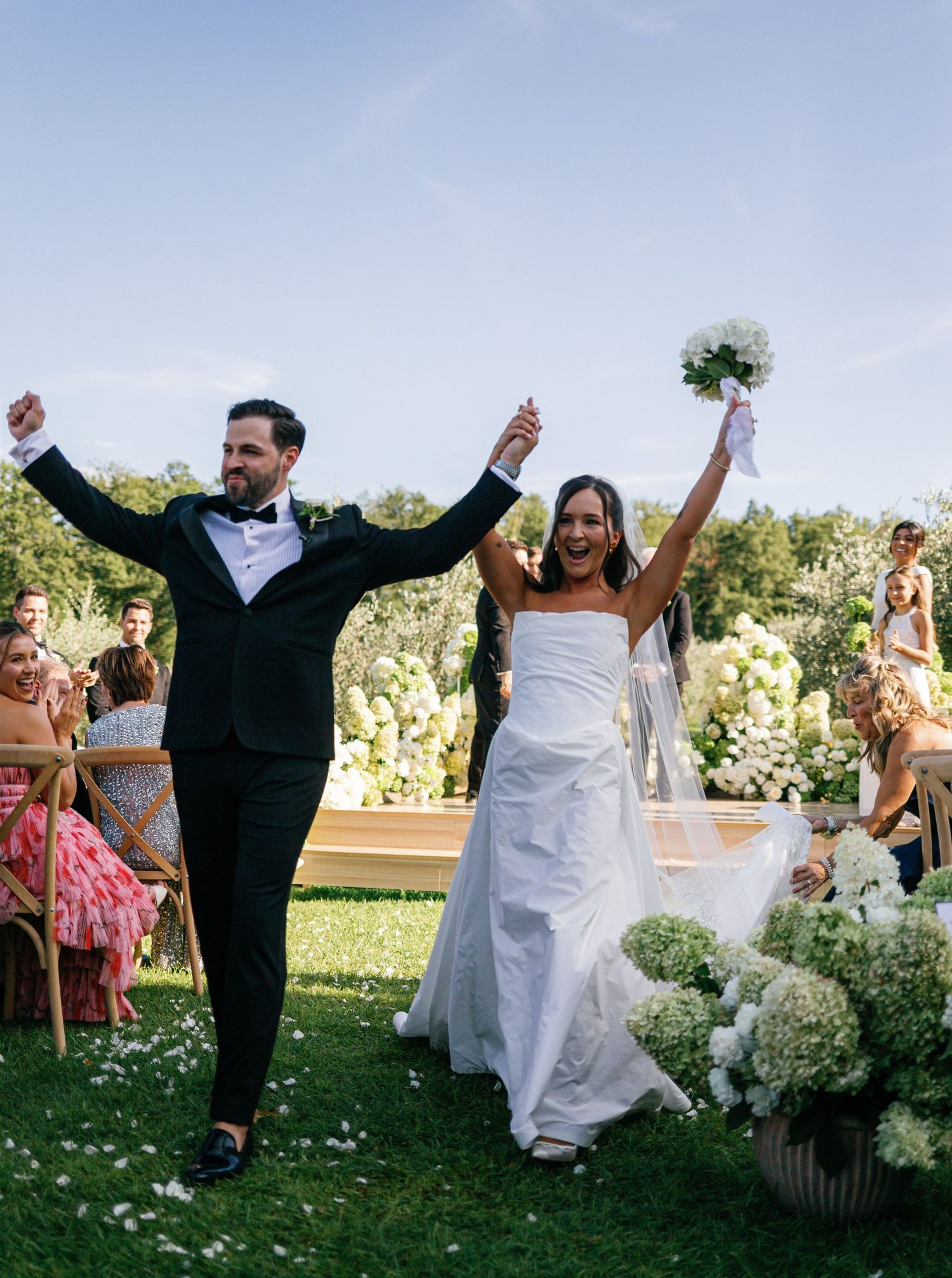 Joyful couple celebrates during outdoor wedding ceremony with guests throwing flower petals in garden setting.