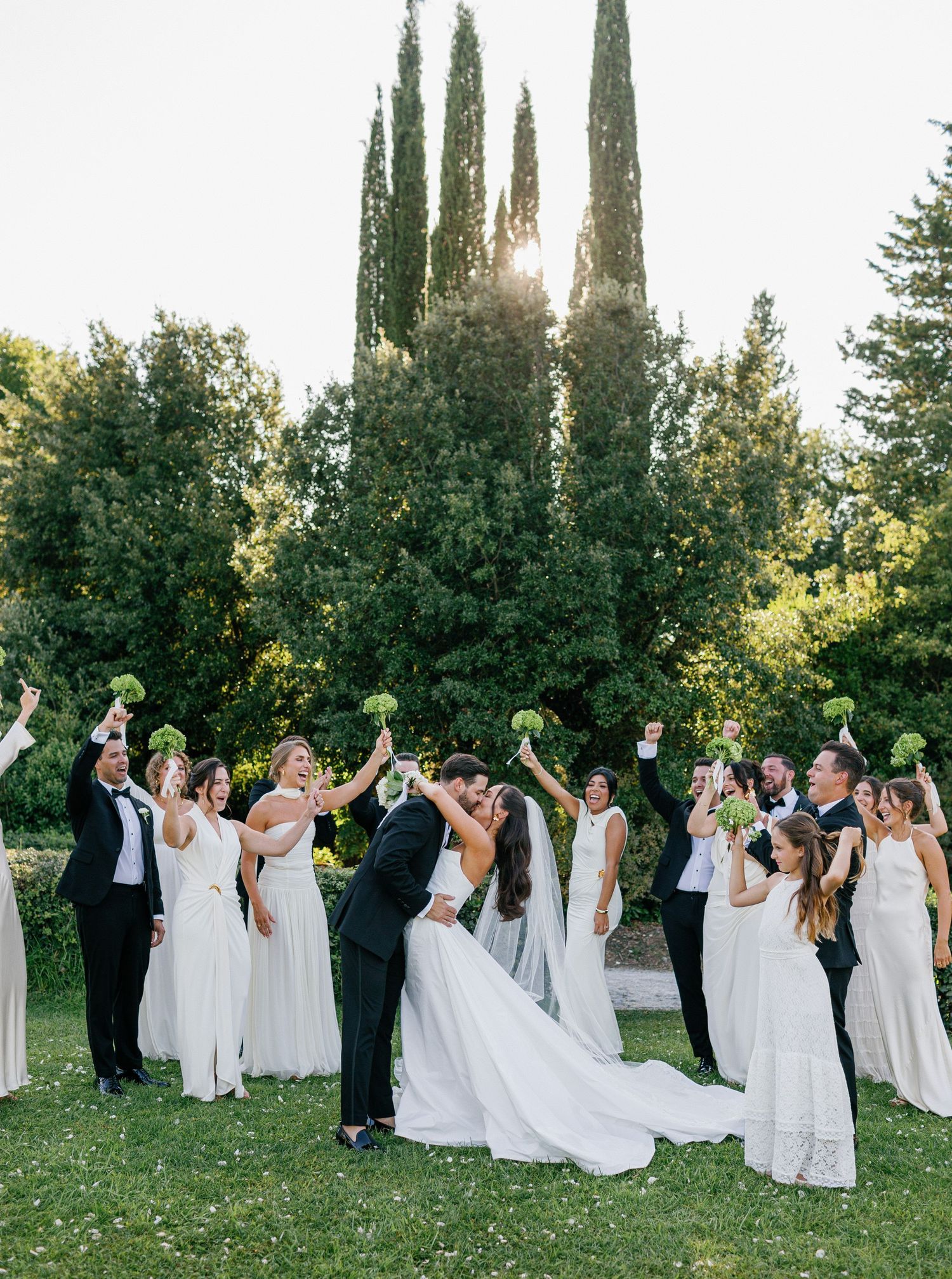 A wedding party in white dresses and formal attire poses together on a lawn with tall cypress trees in the background.
