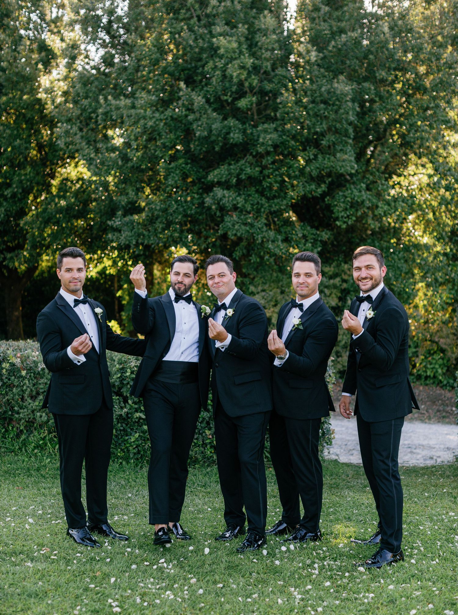 Groomsmen in black tuxedos pose together outdoors with celebratory gestures against a green natural backdrop.