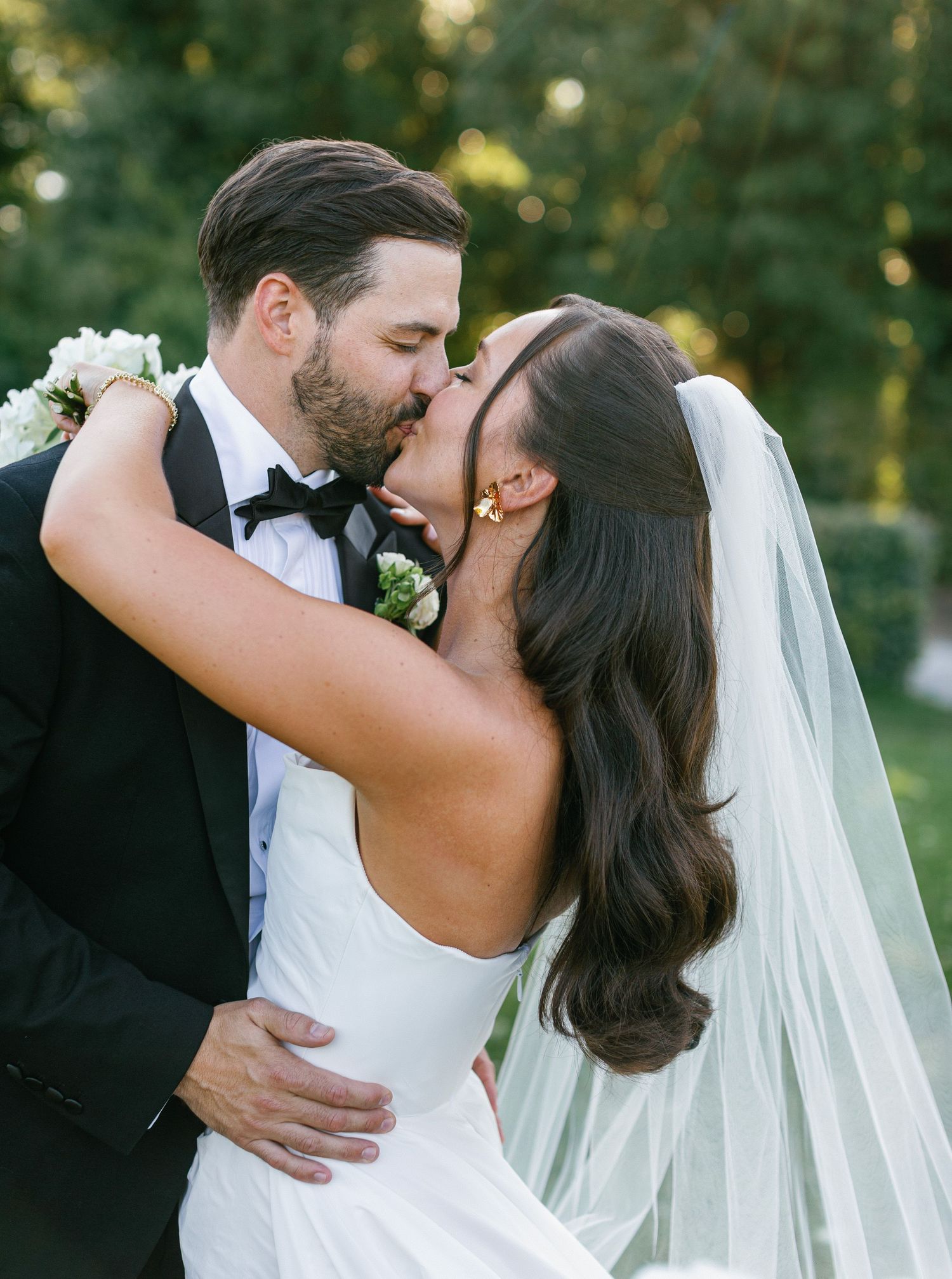 A romantic wedding moment captured outdoors with a flowing white veil and formal attire in natural lighting.