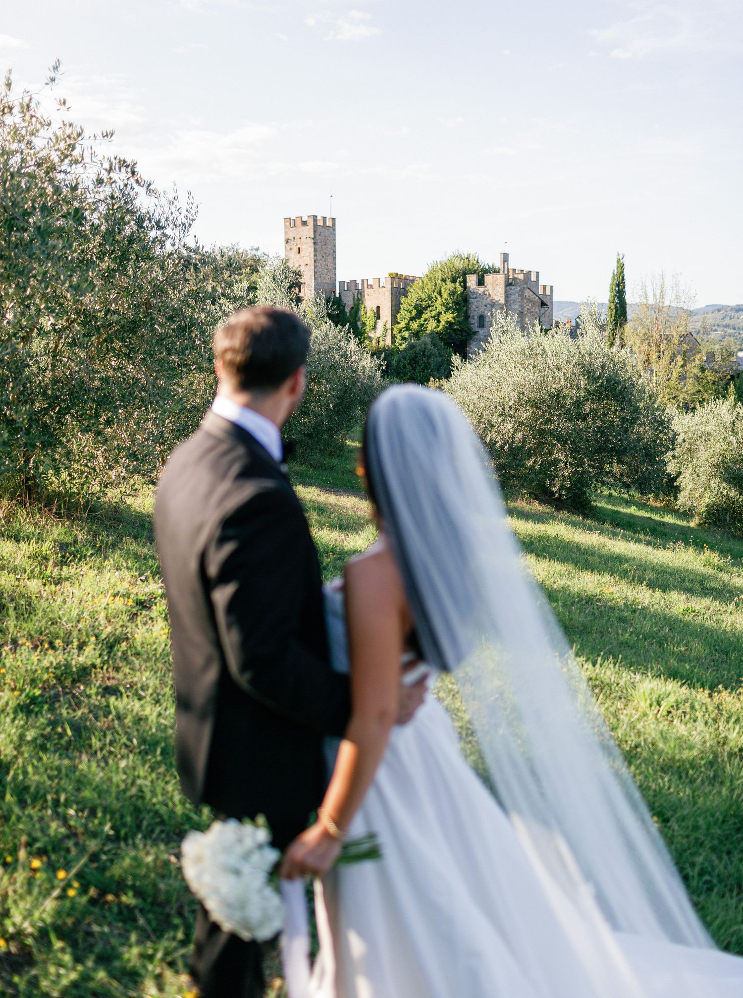 A bride and groom walk together through a lush green garden with stone buildings visible in the background.