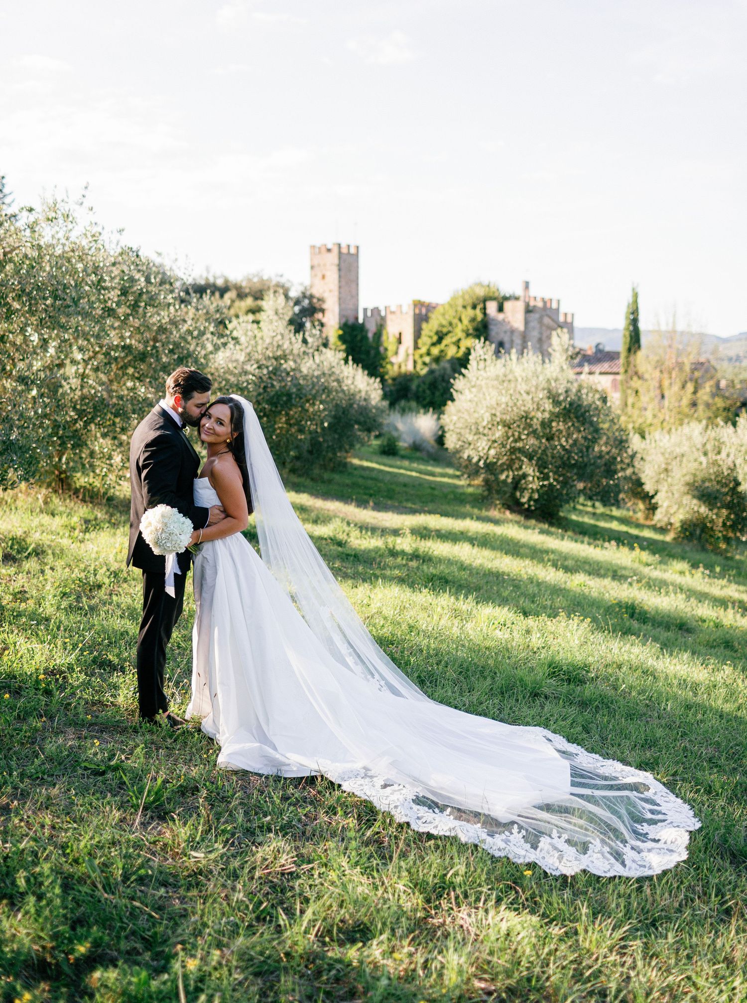 Romantic wedding couple embraces on a sunlit grassy hillside with a castle-like building in the background.