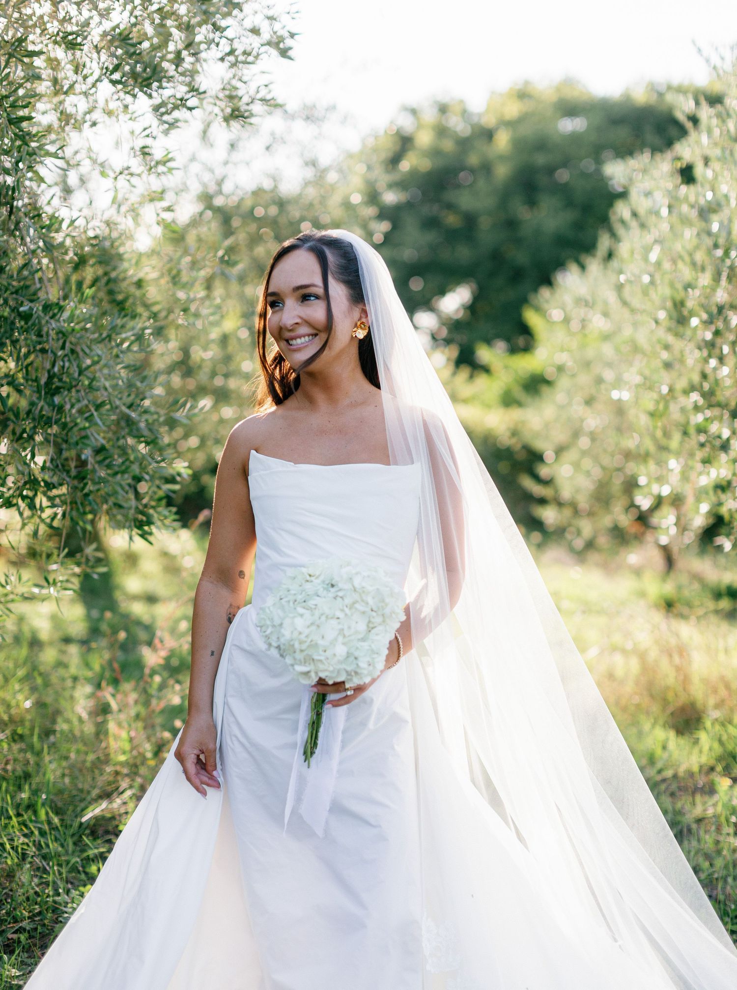 A bride in a classic white strapless wedding gown and long veil holds a white bouquet in a sunlit garden setting.