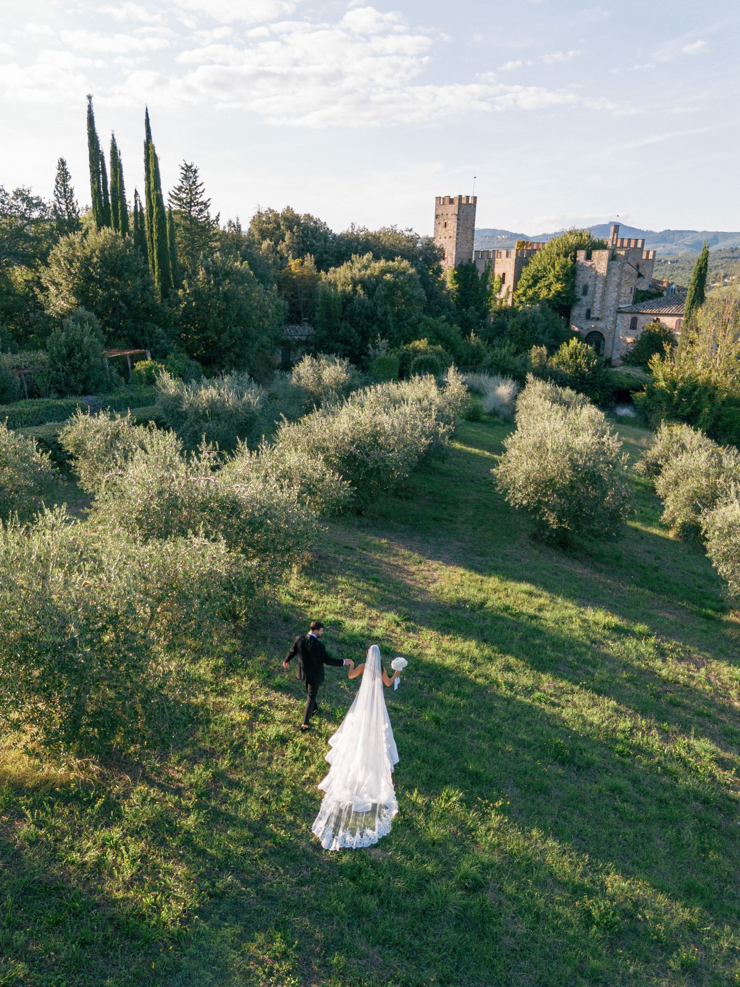 A scenic Tuscan landscape with cypress trees and rolling hills showcases a couple in wedding attire amidst olive groves.