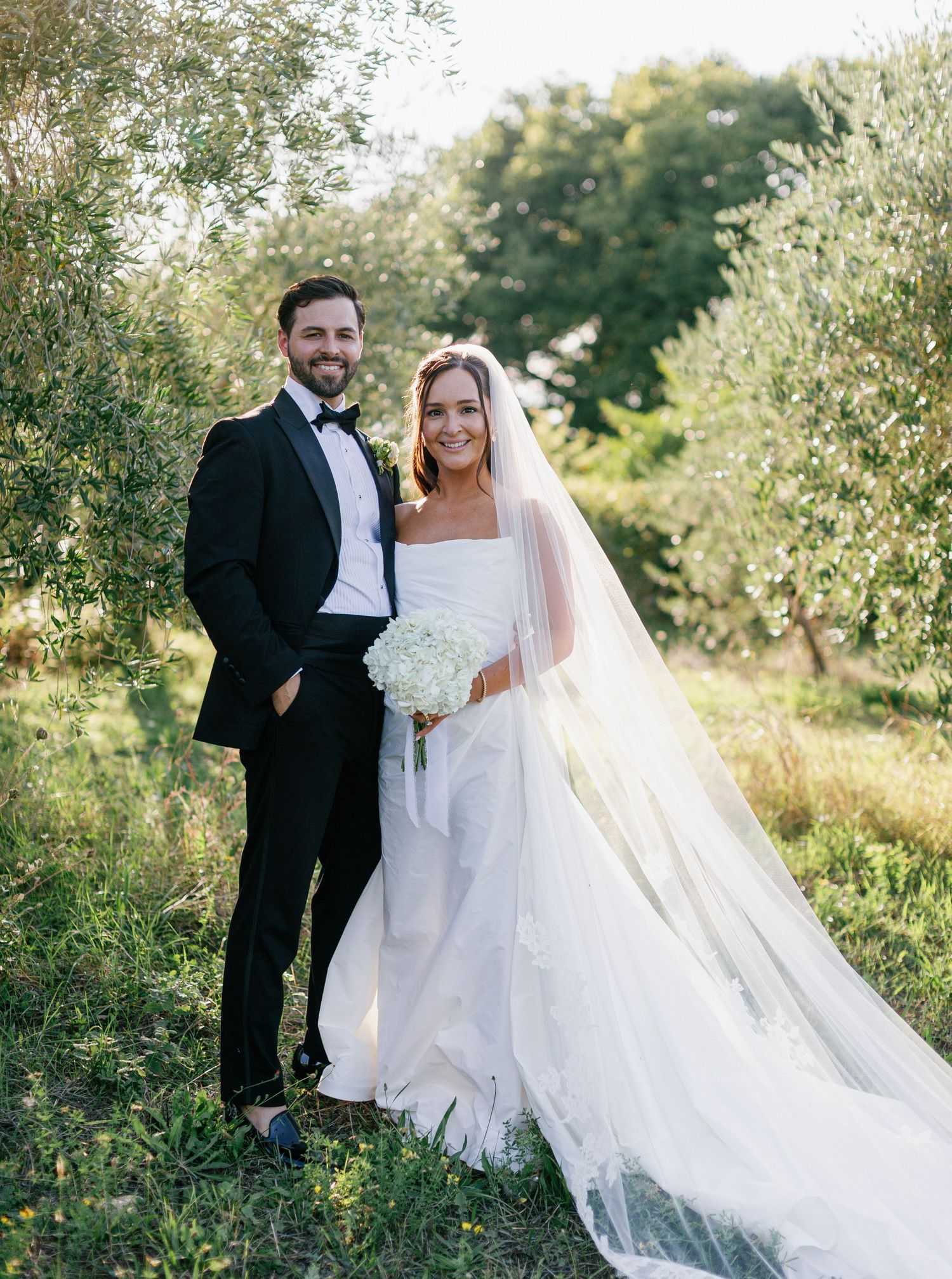 A couple in wedding attire poses together in a sunlit garden setting with flowing white dress and veil.