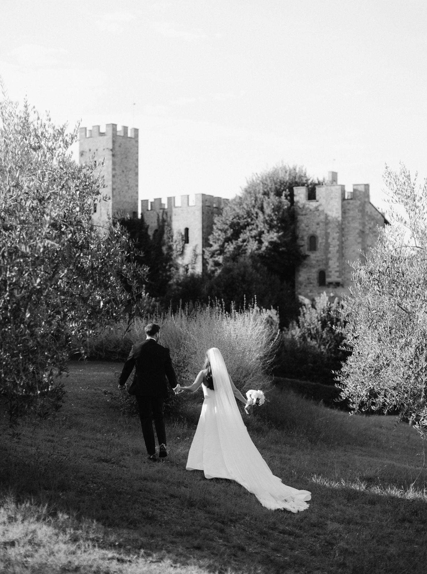 A couple walks along a garden path towards a majestic castle with stone towers and turrets in black and white.