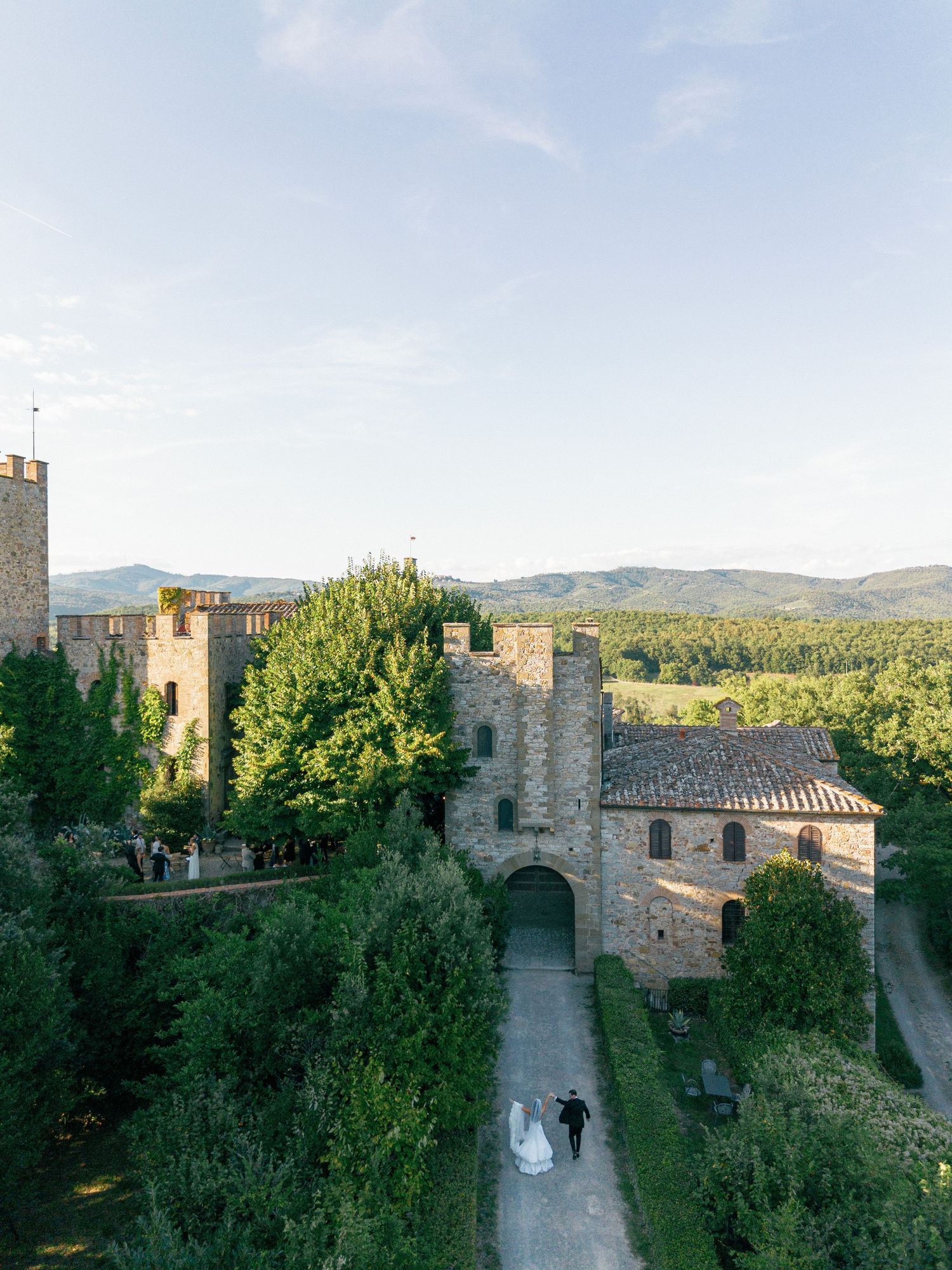 Panoramic view of a medieval stone castle with towers and walkways surrounded by lush green trees in the countryside.