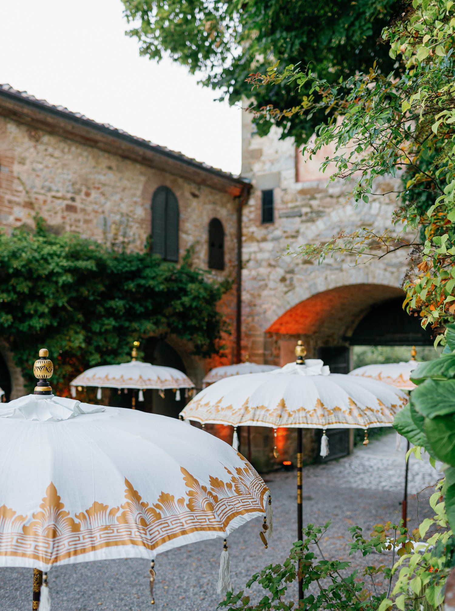 White decorative umbrellas with gold patterns line a rustic stone courtyard in Tuscany at sunset.