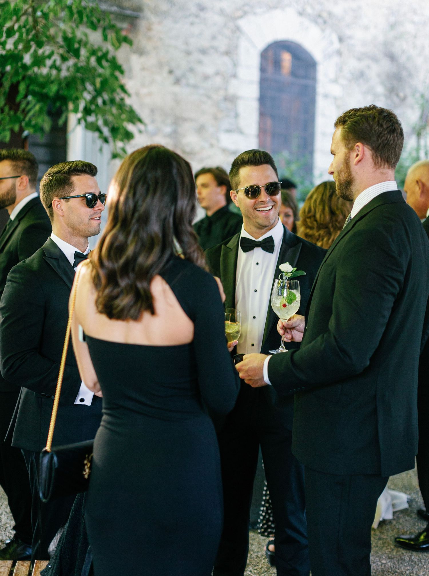 Elegant wedding guests in formal black attire mingle and socialize at an outdoor cocktail reception.