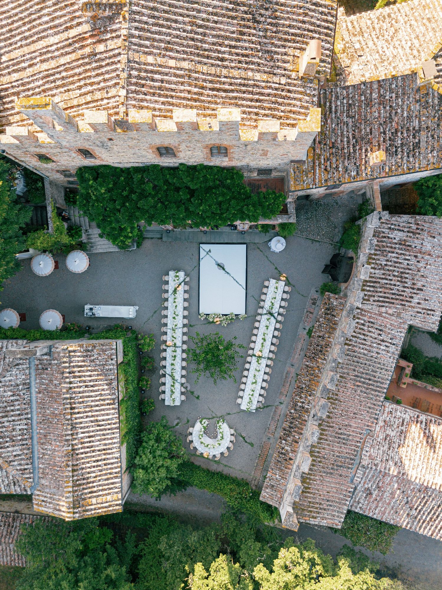 Aerial view of an outdoor wedding reception setup with long tables arranged in a courtyard surrounded by terracotta roofs.