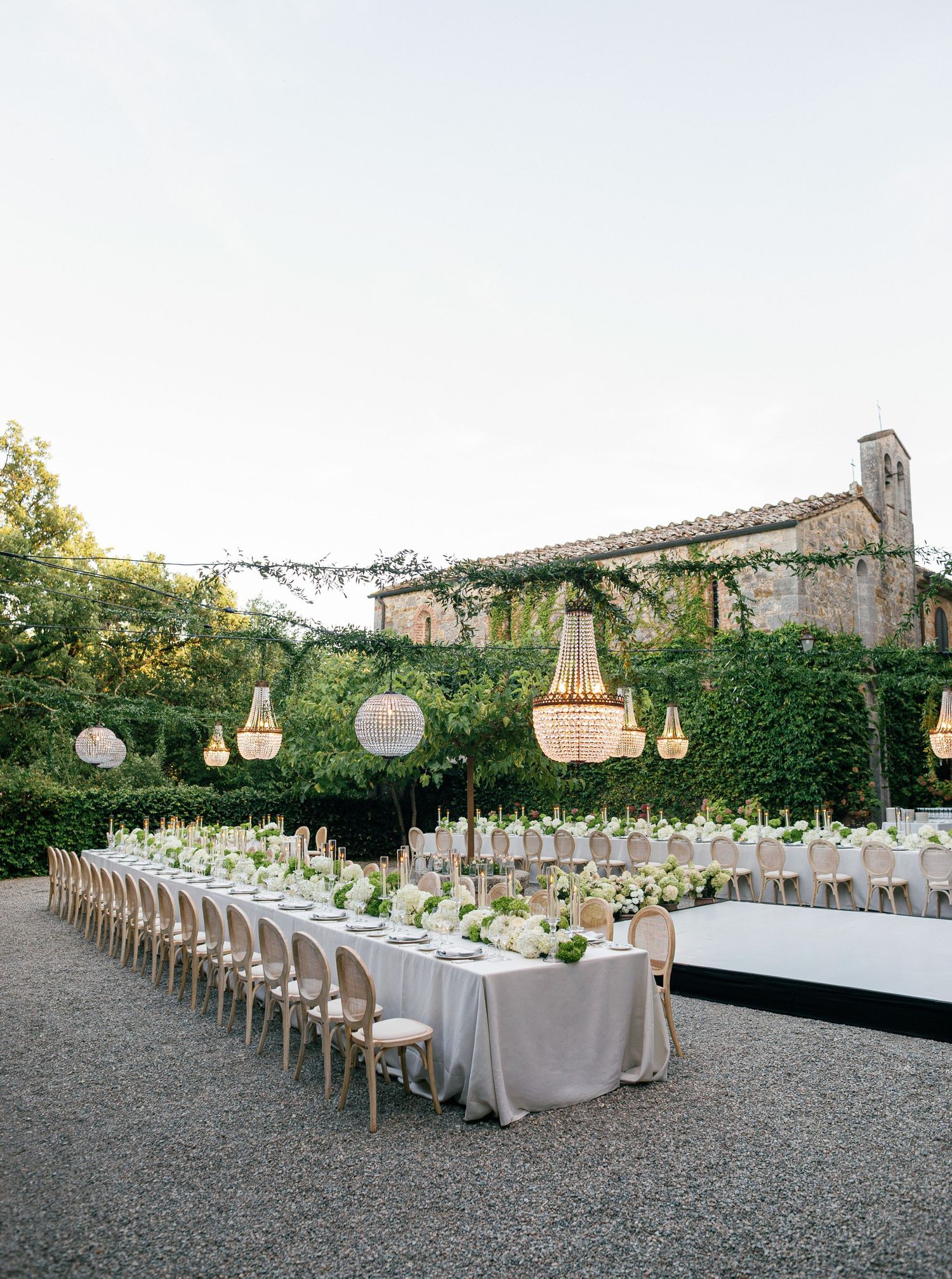 Elegant outdoor wedding reception setup with long tables, crystal chandeliers and lush garden backdrop.