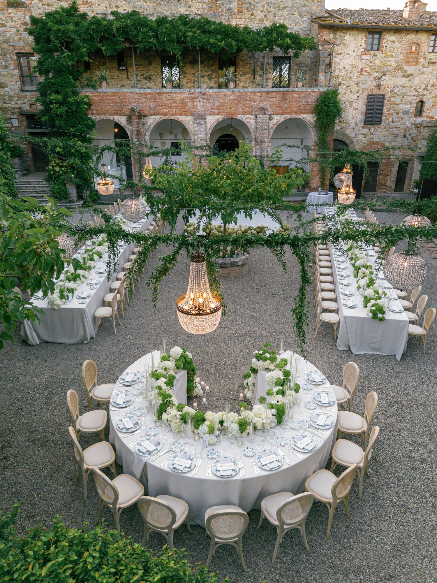 Elegant outdoor wedding reception setup with round tables and natural greenery decor in a rustic Italian courtyard setting.