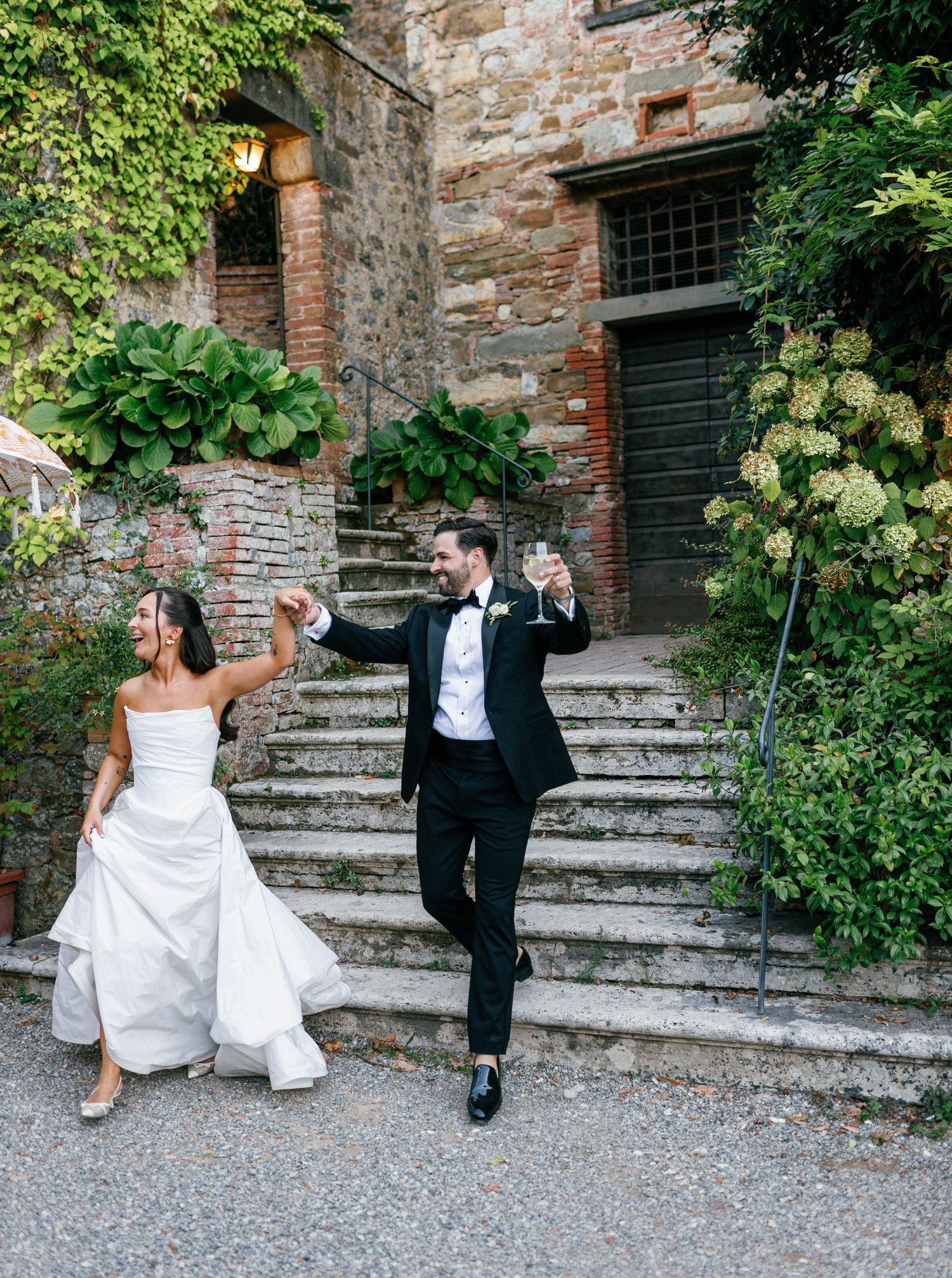 Wedding couple sits playfully on stone steps surrounded by lush greenery at a rustic venue.