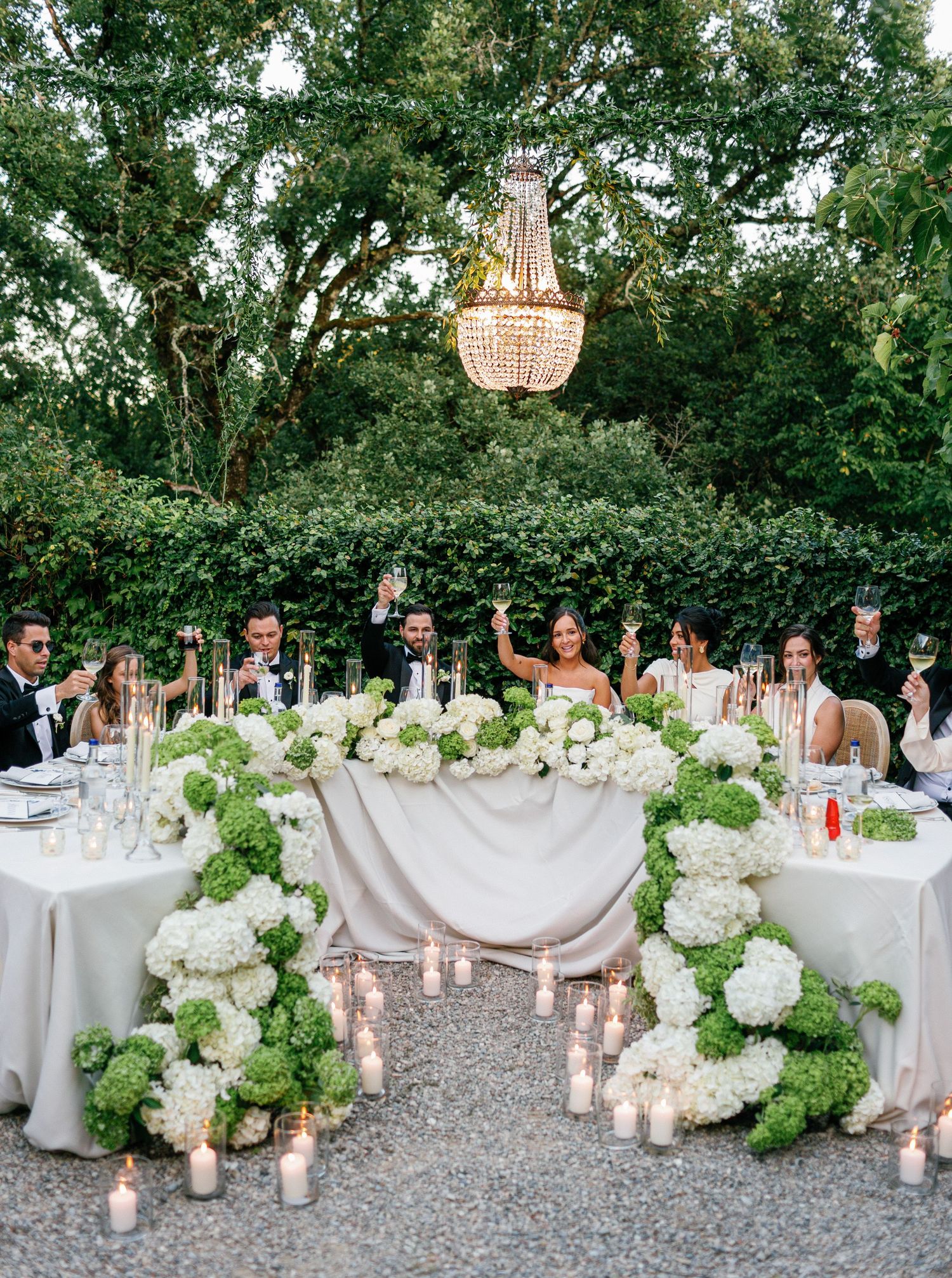 Elegant outdoor wedding reception table decorated with white hydrangeas and candlelight under trees at dusk.